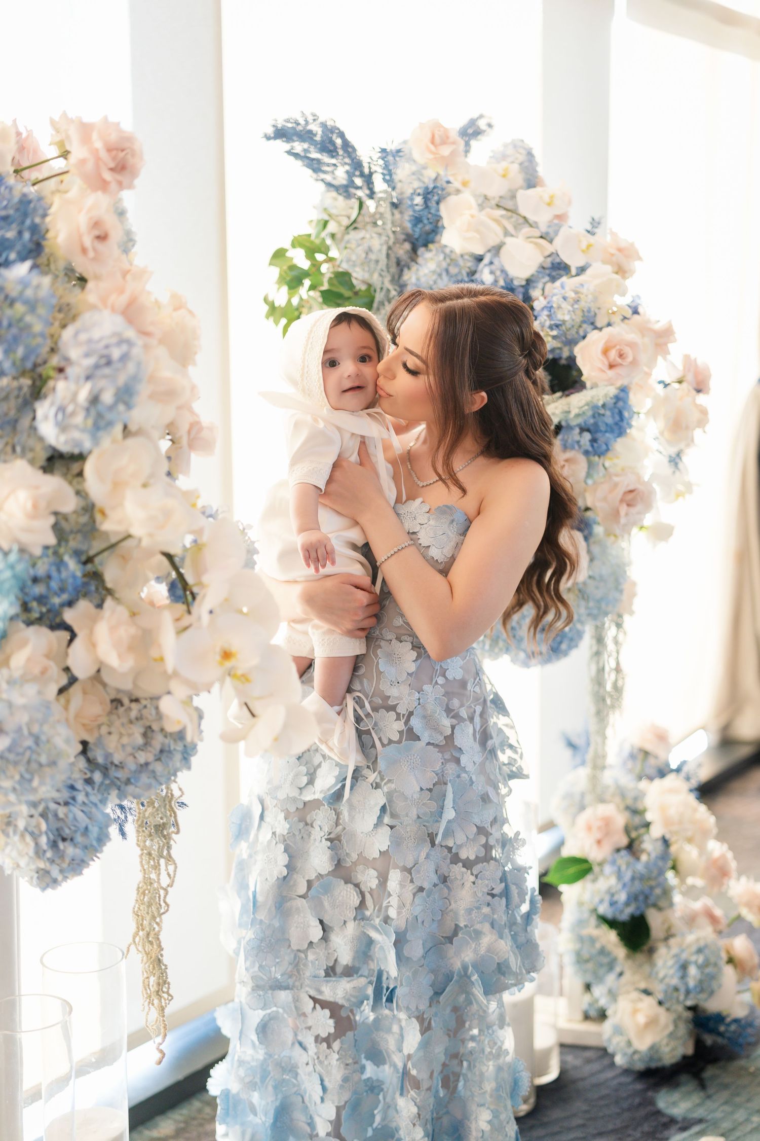 A woman in a flowing blue floral dress holds a baby against a backdrop of blue and white hydrangea flower arrangements.