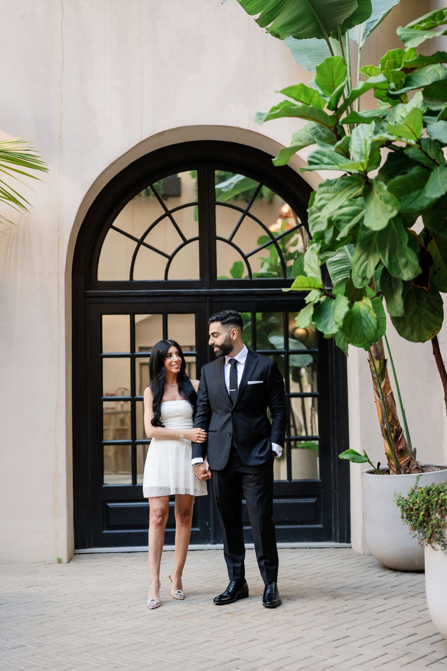 Couple walking together through Mediterranean-style courtyard with large fiddle leaf plants.