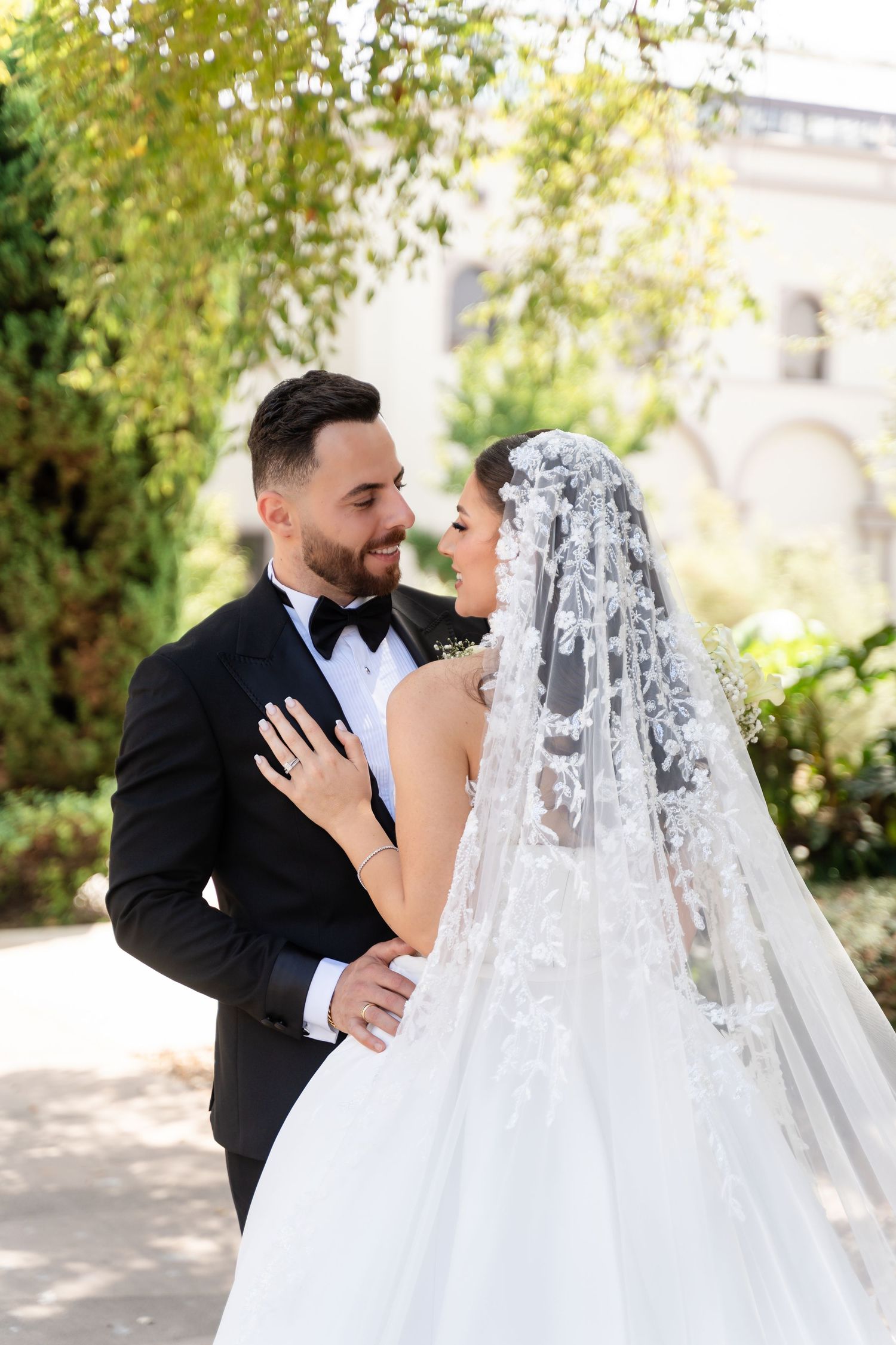 An elegant wedding couple shares an intimate moment under trees in a sunlit garden setting.