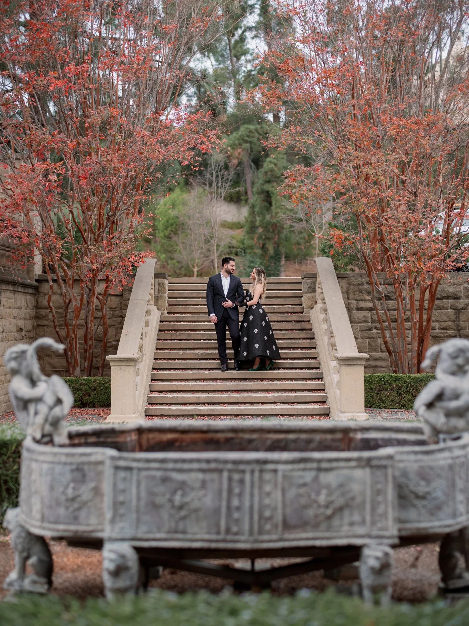 A couple stands on grand stone steps framed by blooming coral trees and classical statuary.