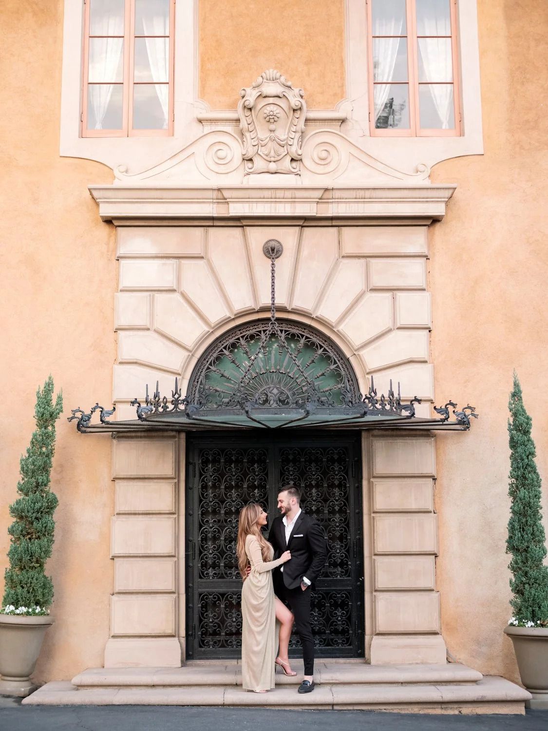 Mediterranean style building facade with ornate ironwork and cypress trees in decorative planters.