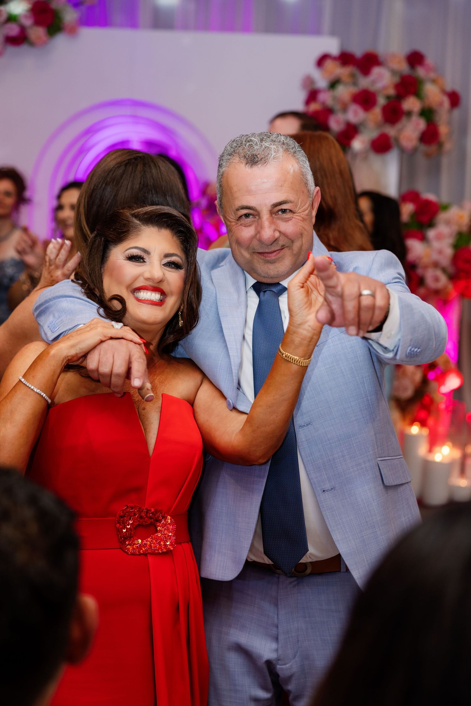 Couple in elegant attire smiling and pointing at a festive wedding reception with floral decorations.