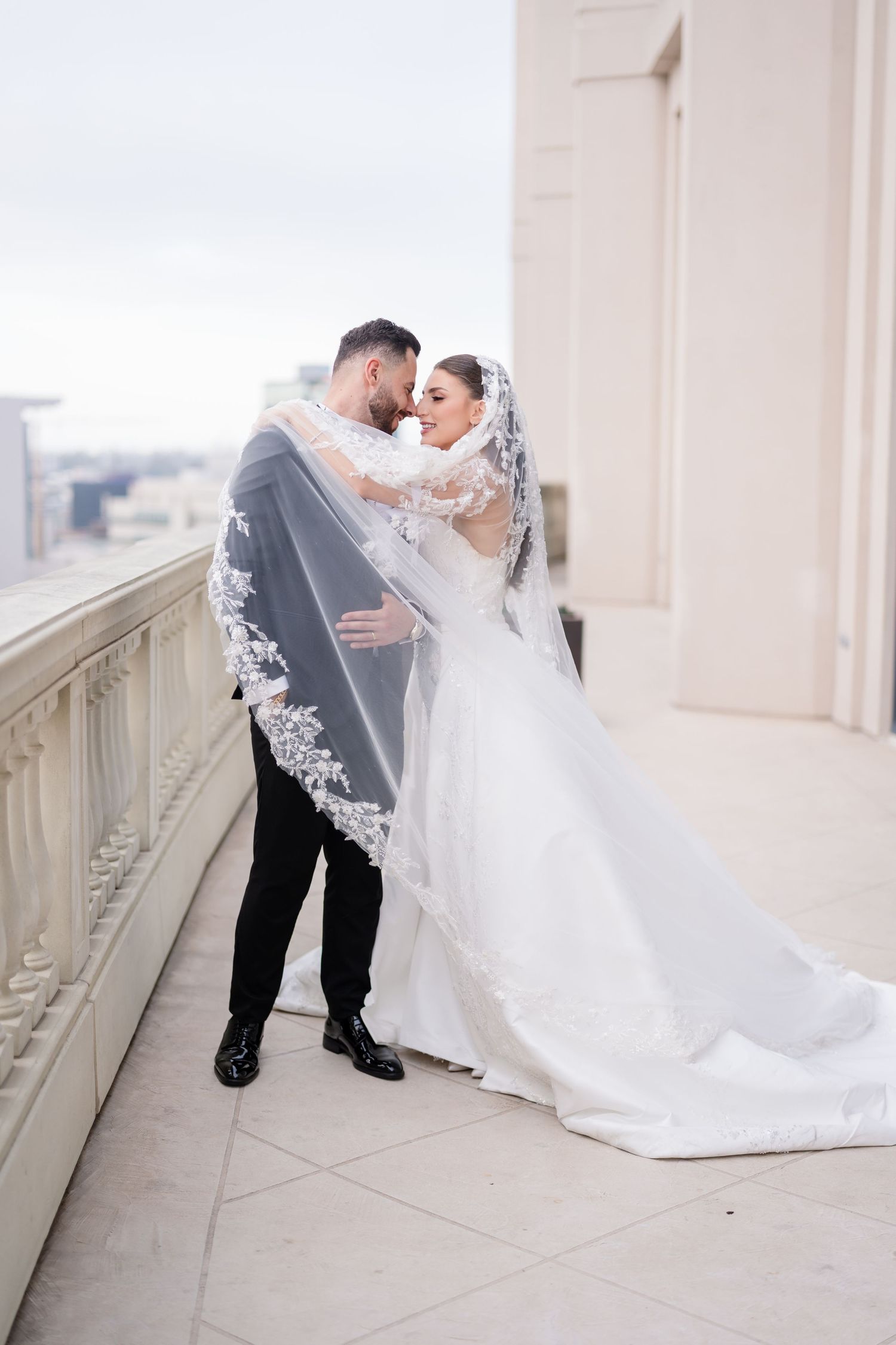 Romantic wedding couple embracing on an outdoor balcony with a flowing white veil.
