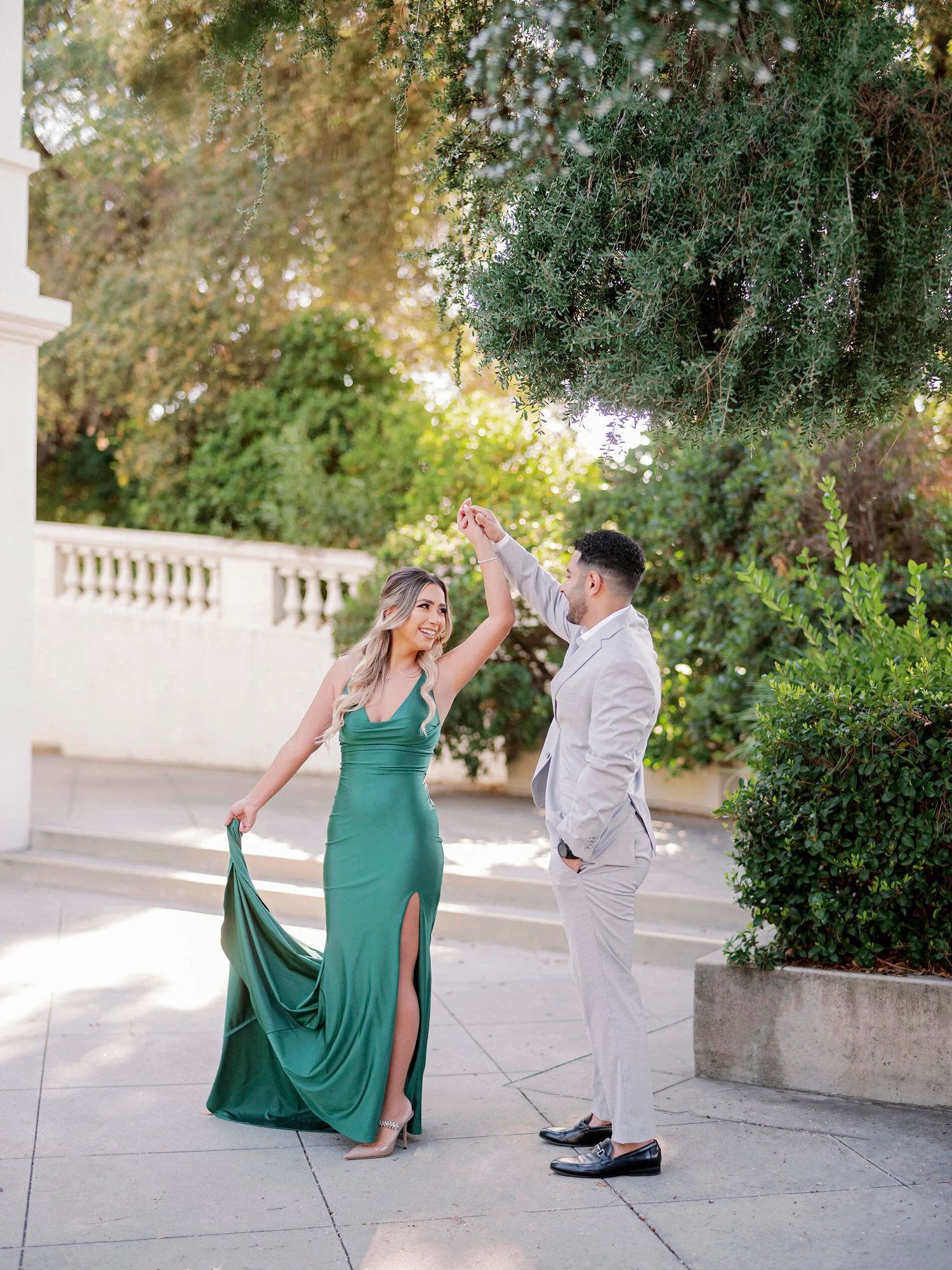 A couple in formal attire dances elegantly on a white stone terrace surrounded by lush greenery.