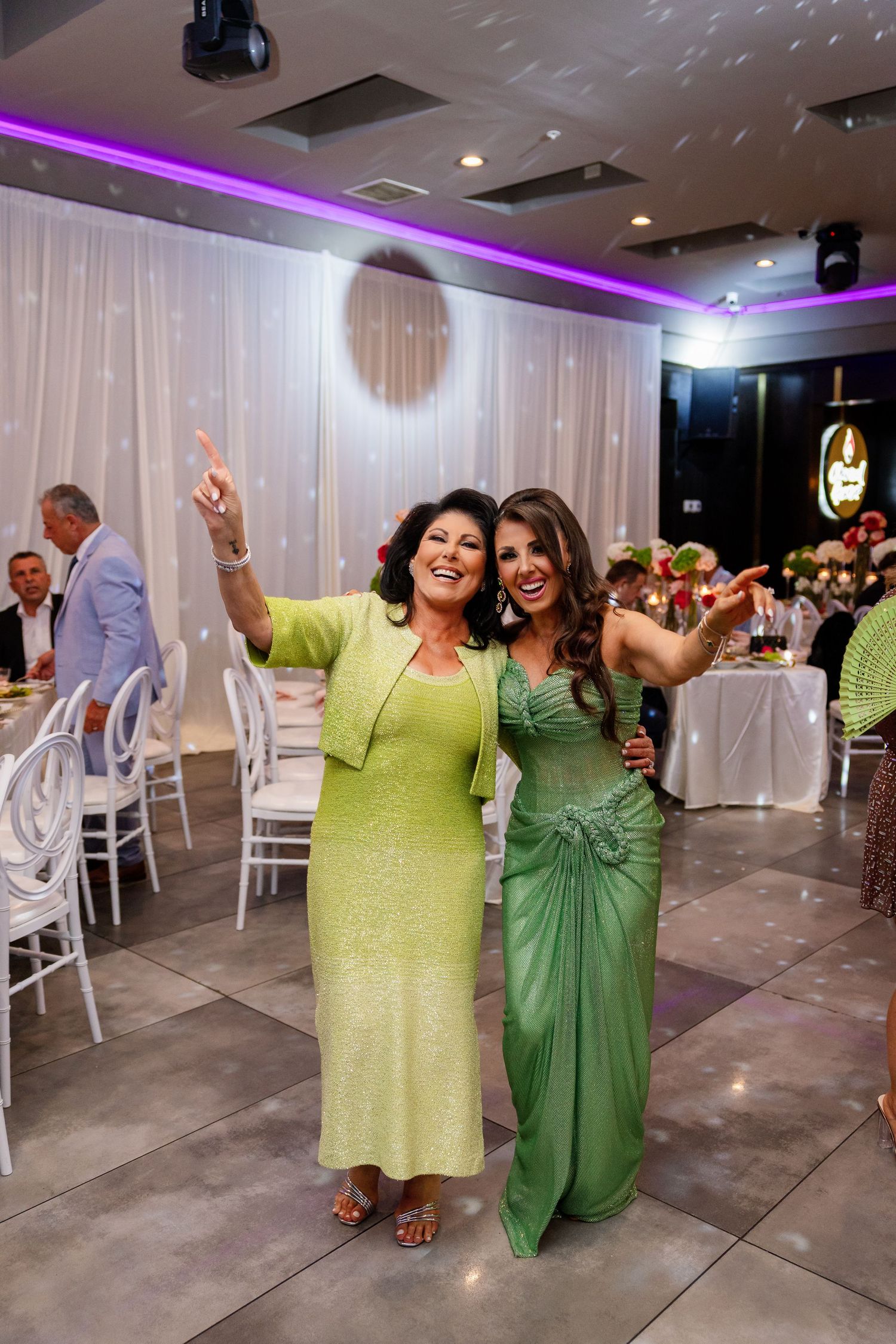 Two women in green dresses dancing and pointing up at a wedding reception venue with purple lighting.