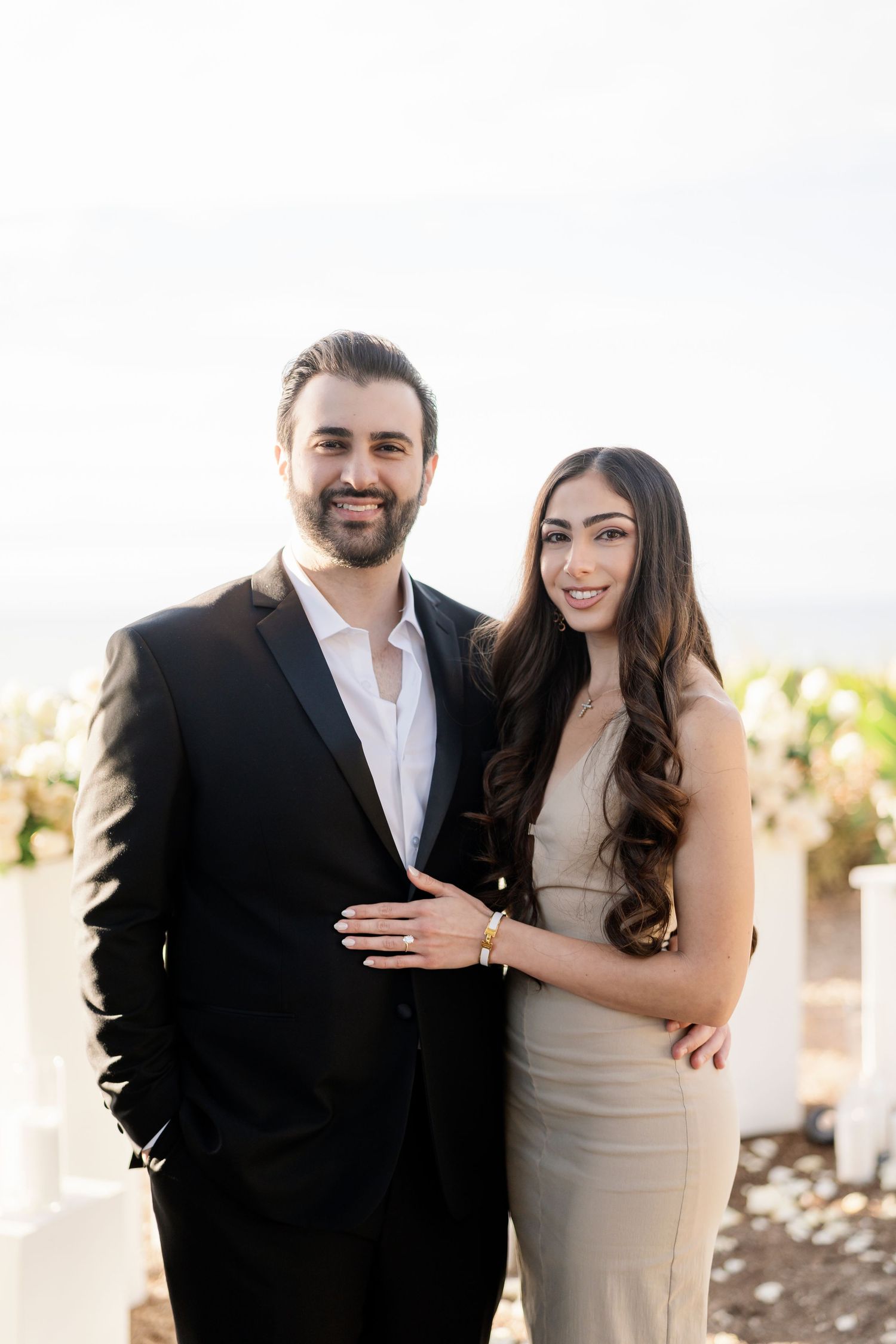 A couple poses for engagement photos in formal attire with a light background.