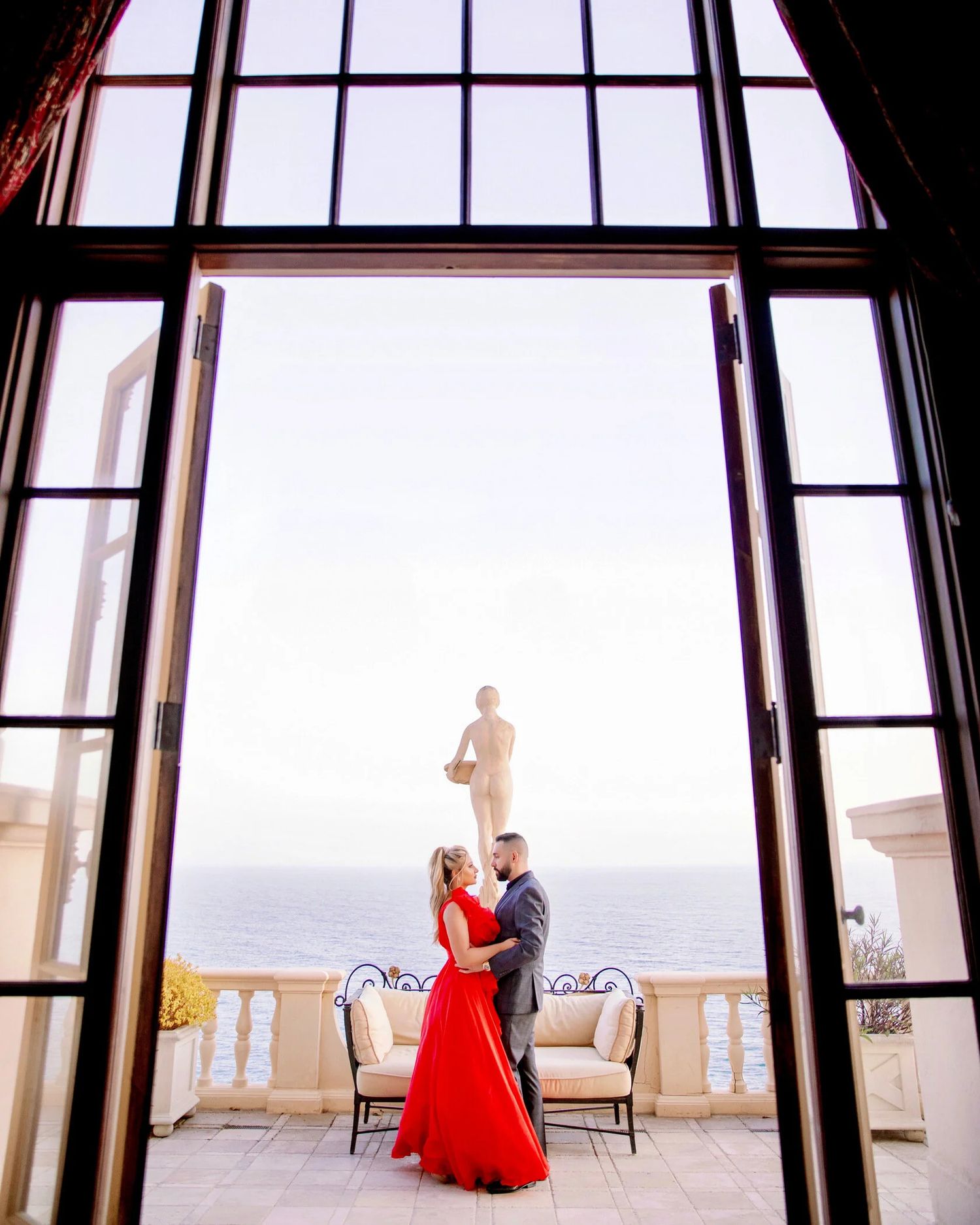 A couple embraces by large windows overlooking a balcony with bright natural light streaming in.