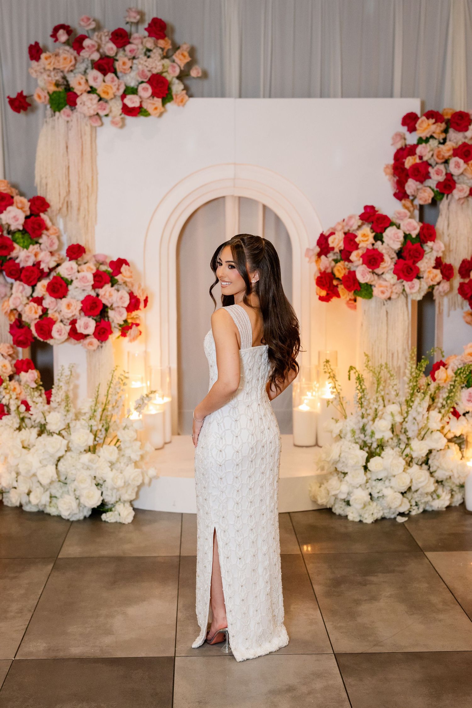 Woman in white sequin gown poses before floral arch with red and pink roses and white candles.