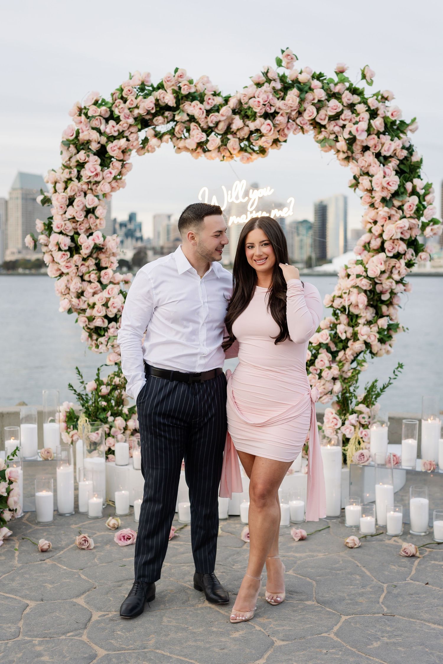 Romantic waterfront proposal scene with pink floral arch and white candles at sunset.
