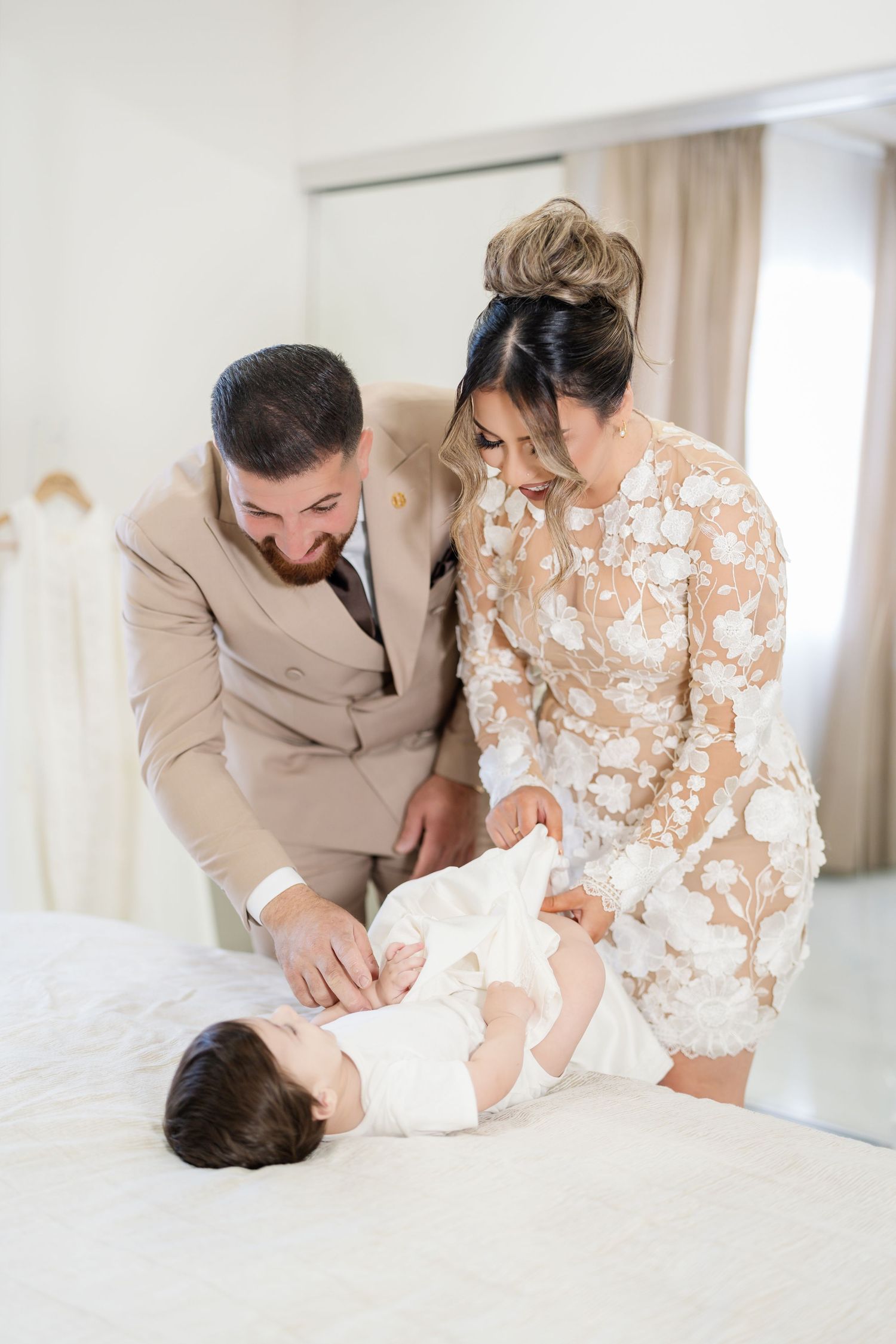 A couple in formal attire shares a tender moment with their baby on a white bed.
