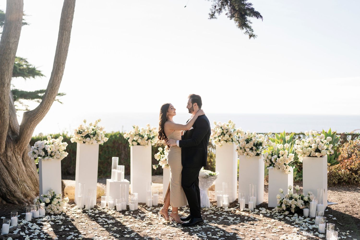 A romantic proposal scene with white floral pillars under forest trees.