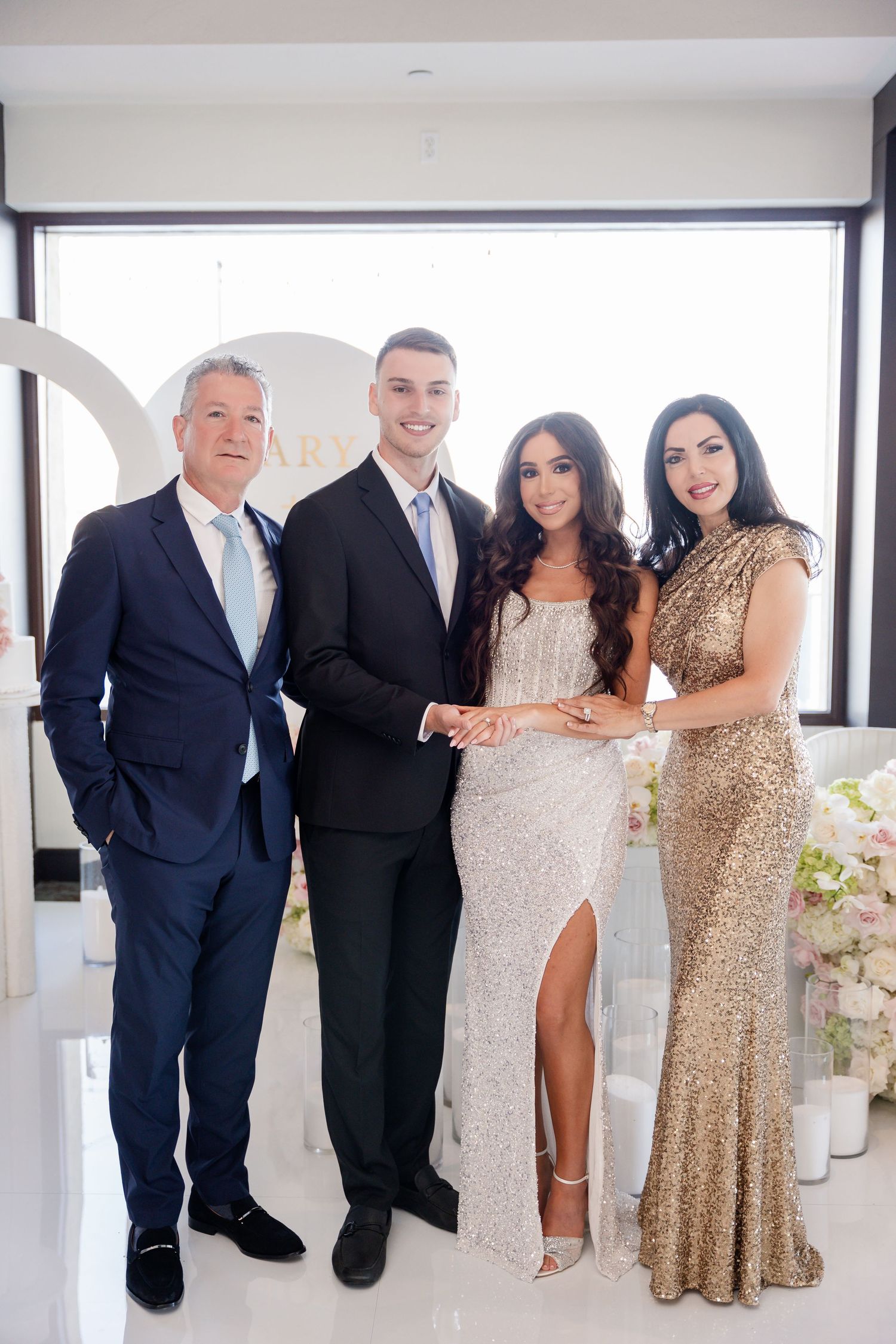 Wedding party poses in formal attire with men in dark suits and women in sparkly gold and white dresses.