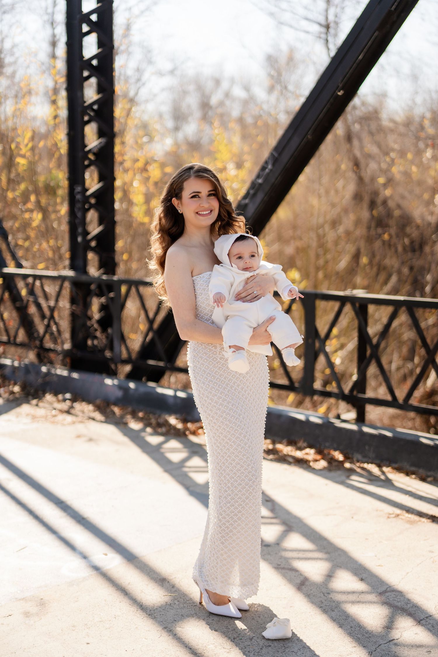 A parent in a white dress holds a baby while standing on a bridge in autumn sunlight.