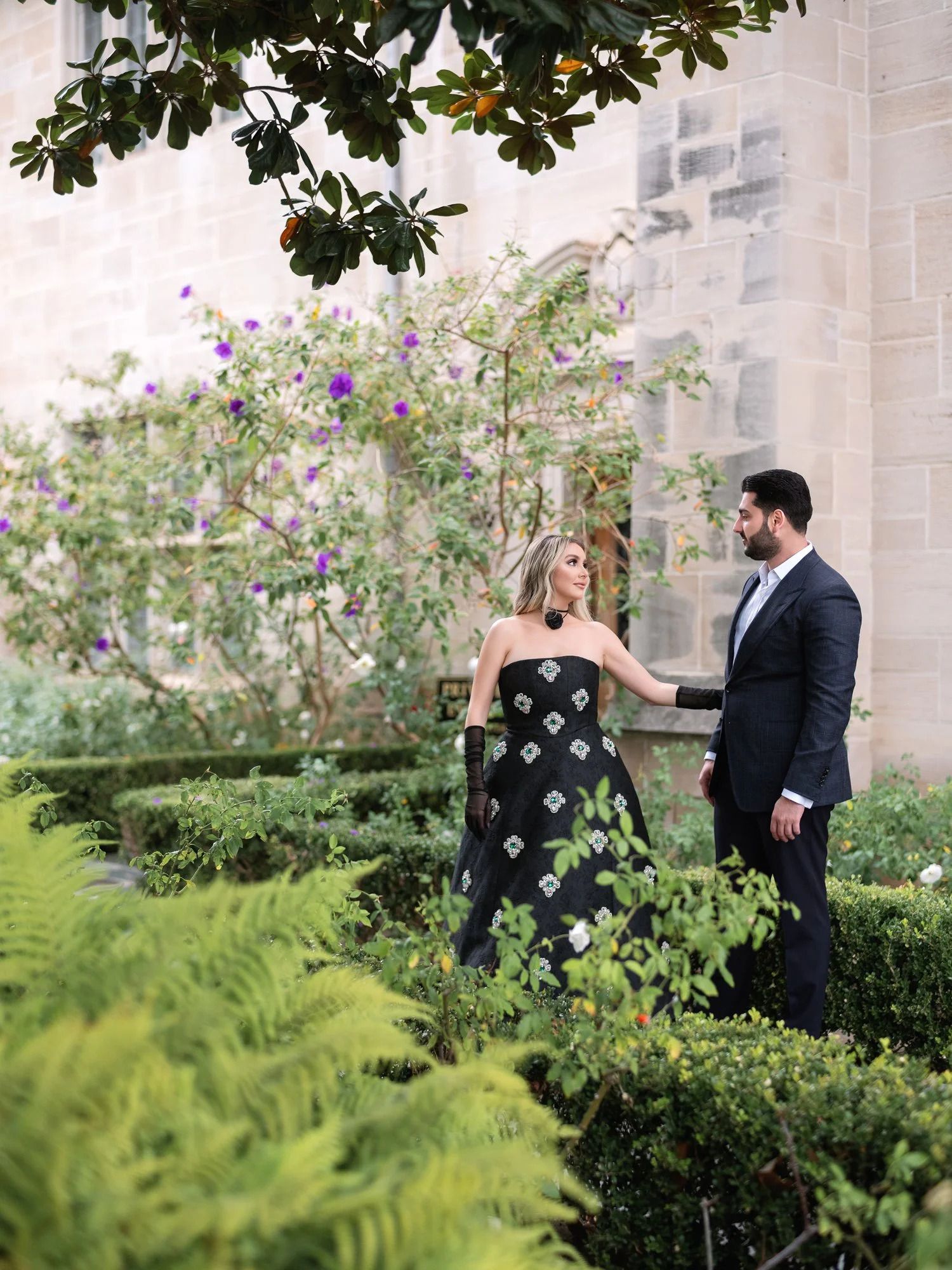 A couple stands in a lush garden setting with blooming flowers and greenery.