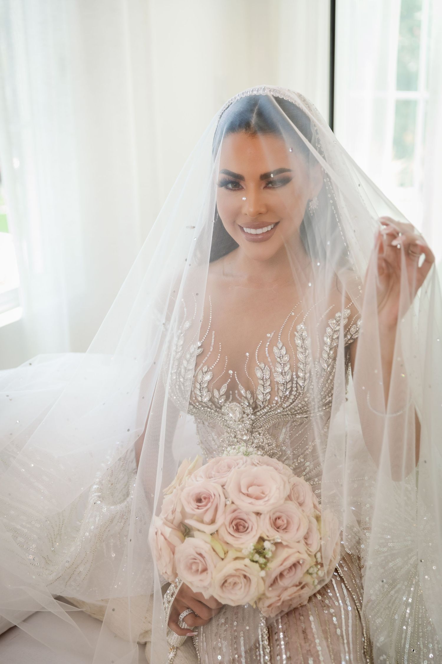 Bride holding a soft pink rose bouquet with a sparkly veil in natural window light.