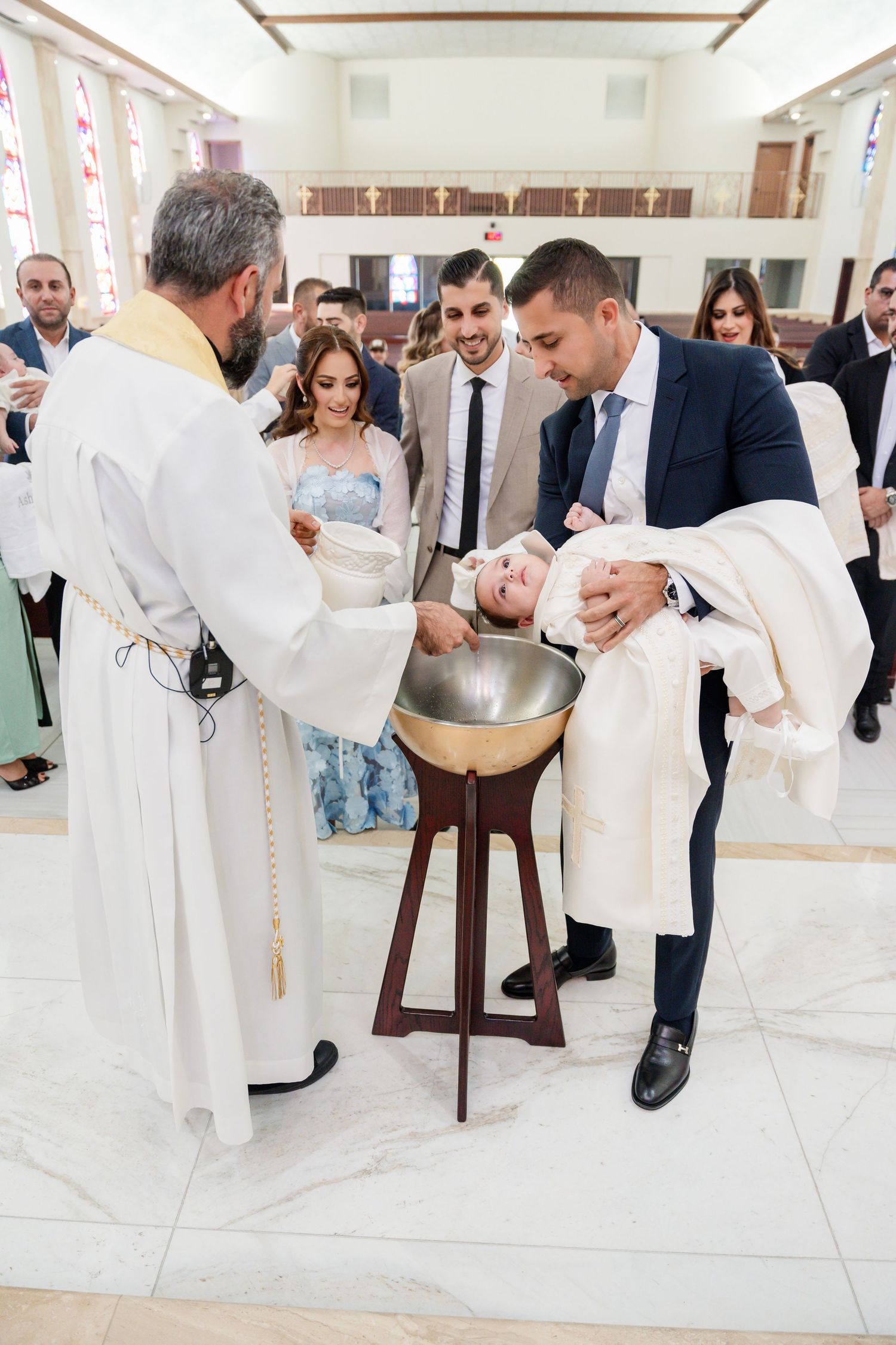 A baptismal ceremony takes place in a church as a priest performs the ritual over a baptismal font on a wooden stand.