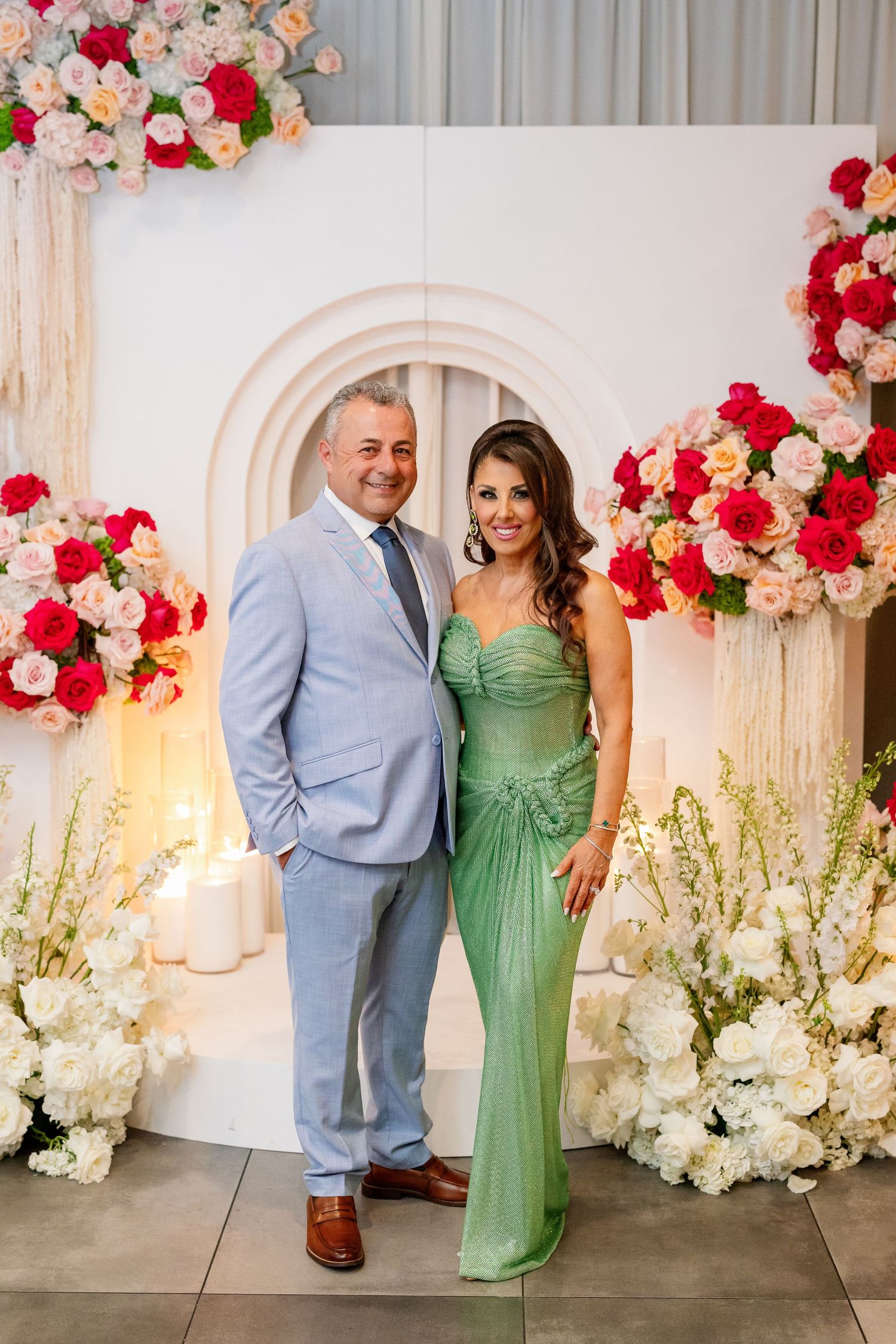 Couple posing at floral backdrop, man in blue suit, woman in green gown, surrounded by pink and white flowers.