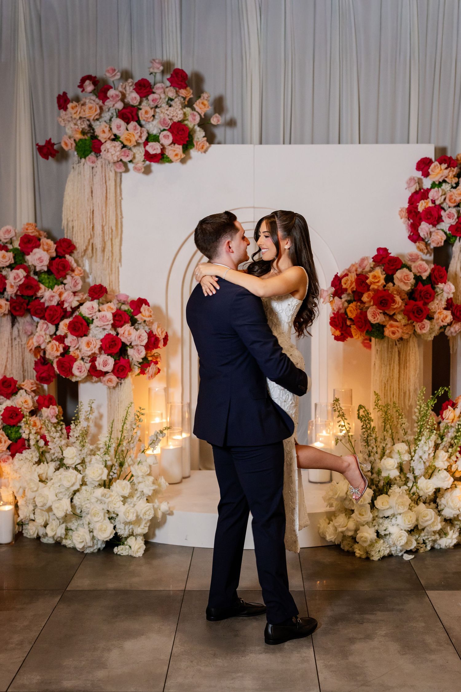 Couple embracing in front of a floral arch with red, pink, and white roses during a romantic proposal setup.