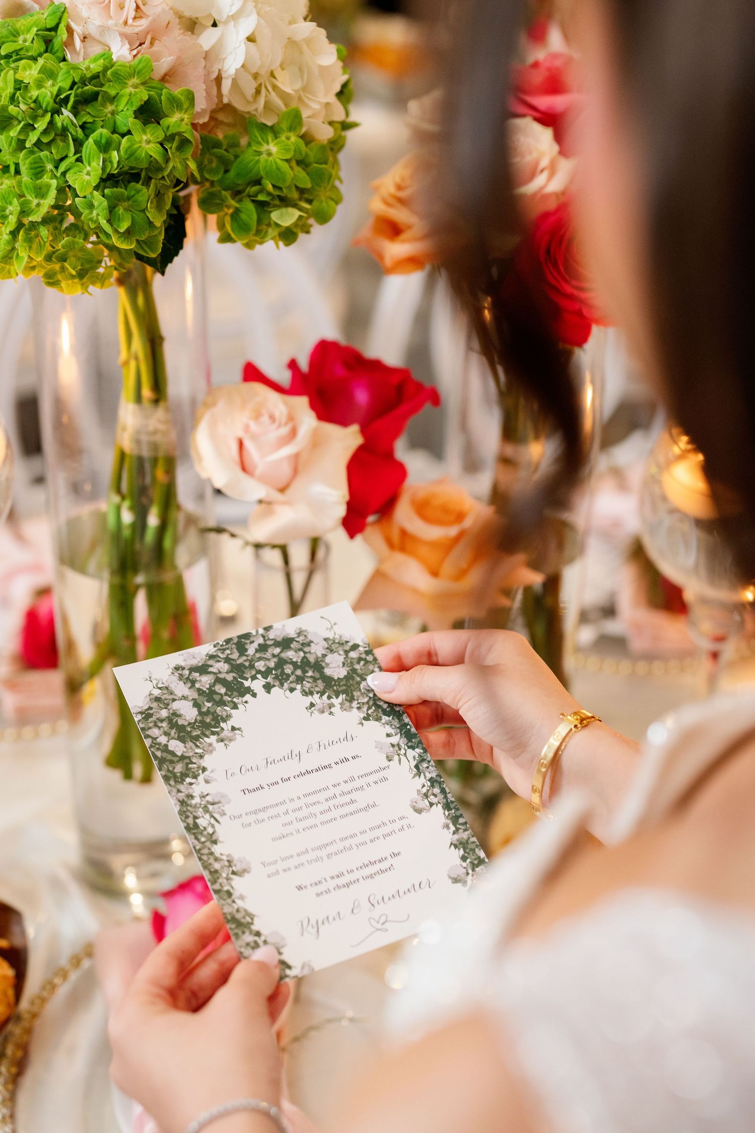 Bride holds elegant floral wedding menu card at reception table with pink, red, and green flower arrangements.