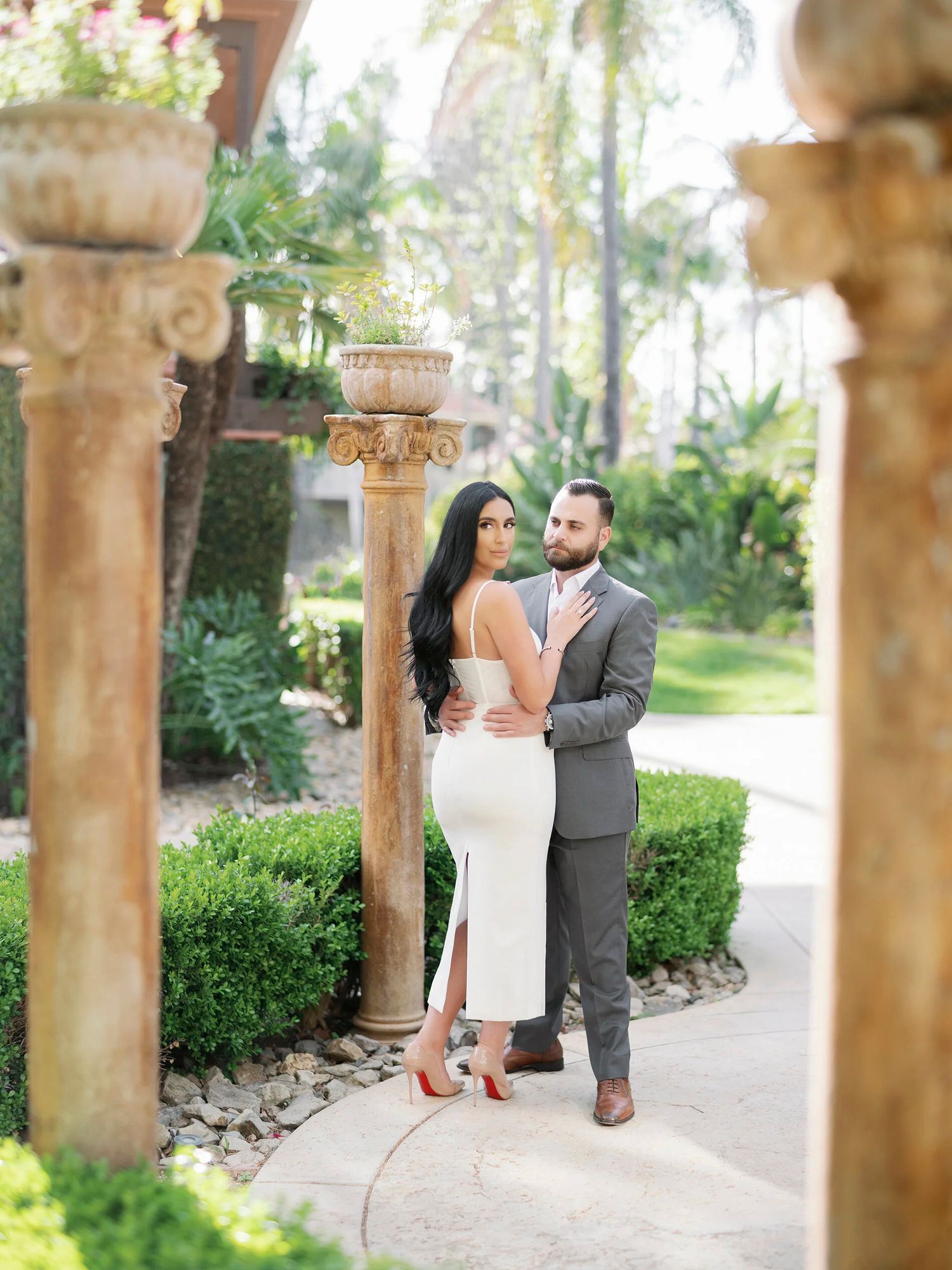 Elegant couple posing between stone columns in a manicured garden walkway.