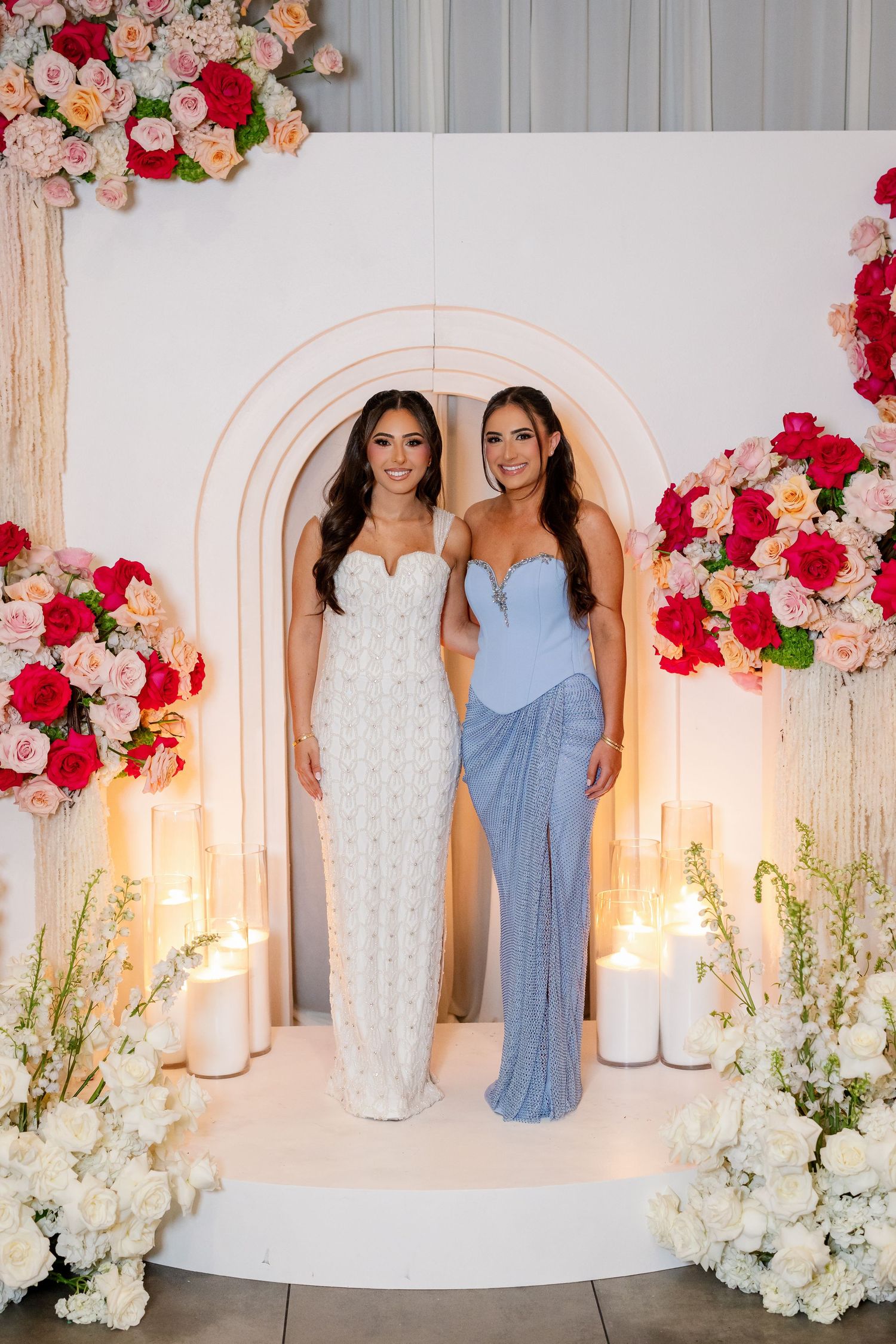 Two women in elegant gowns pose before a floral arch with candles and pink roses at a formal event.