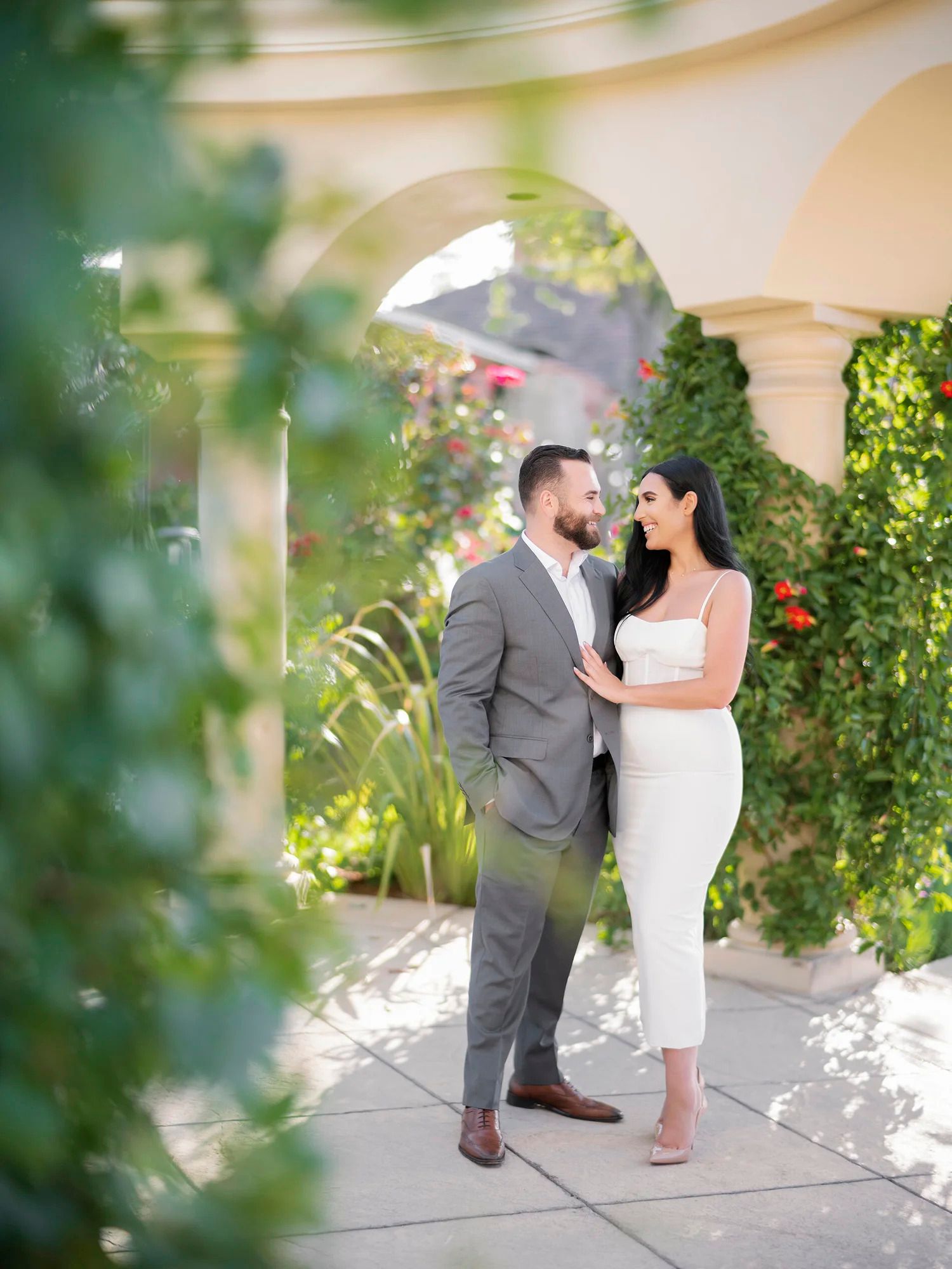 A couple shares an intimate moment in a sunlit garden walkway with blooming flowers.