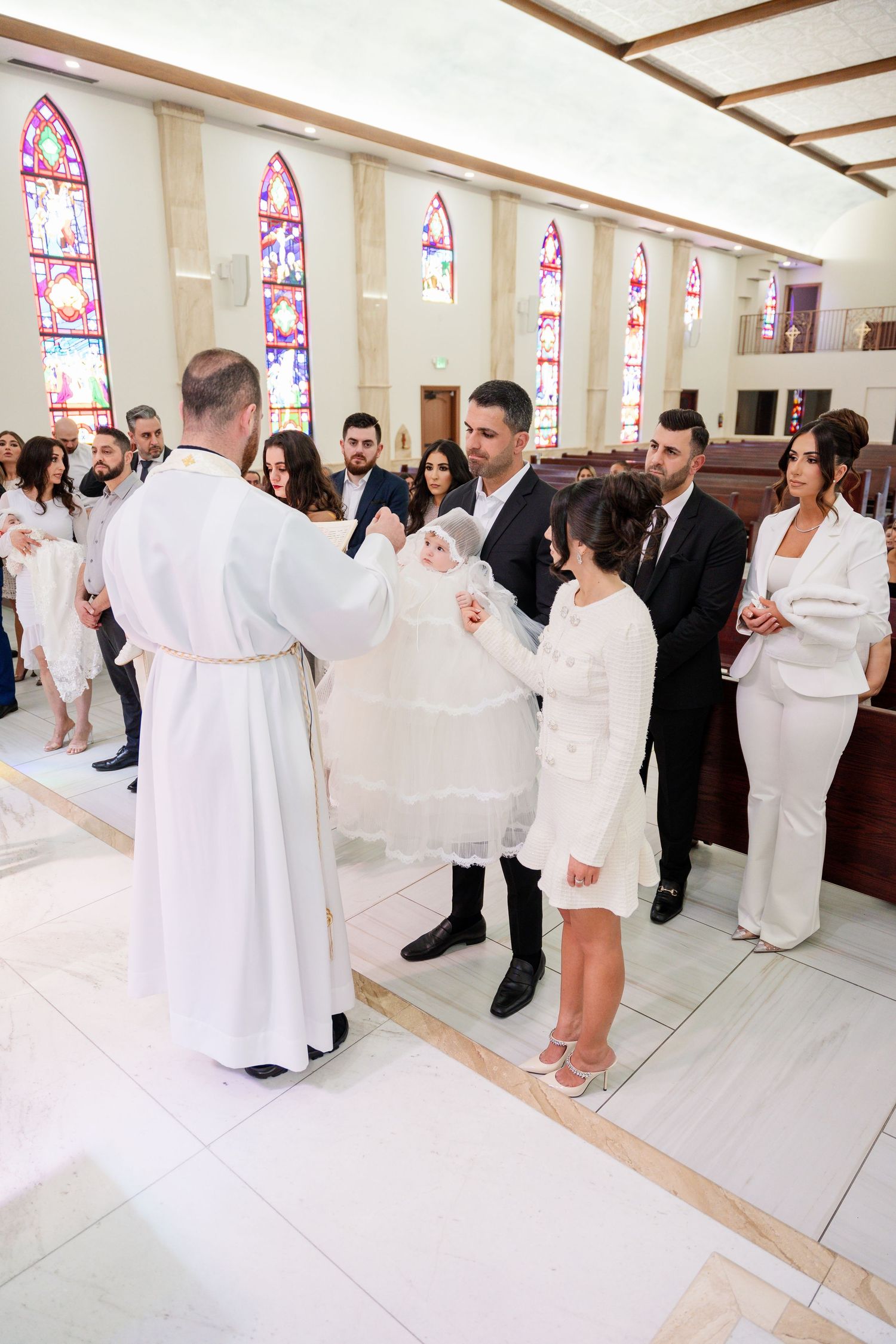 A group of people in white attire participate in a religious ceremony inside a church with stained glass windows.