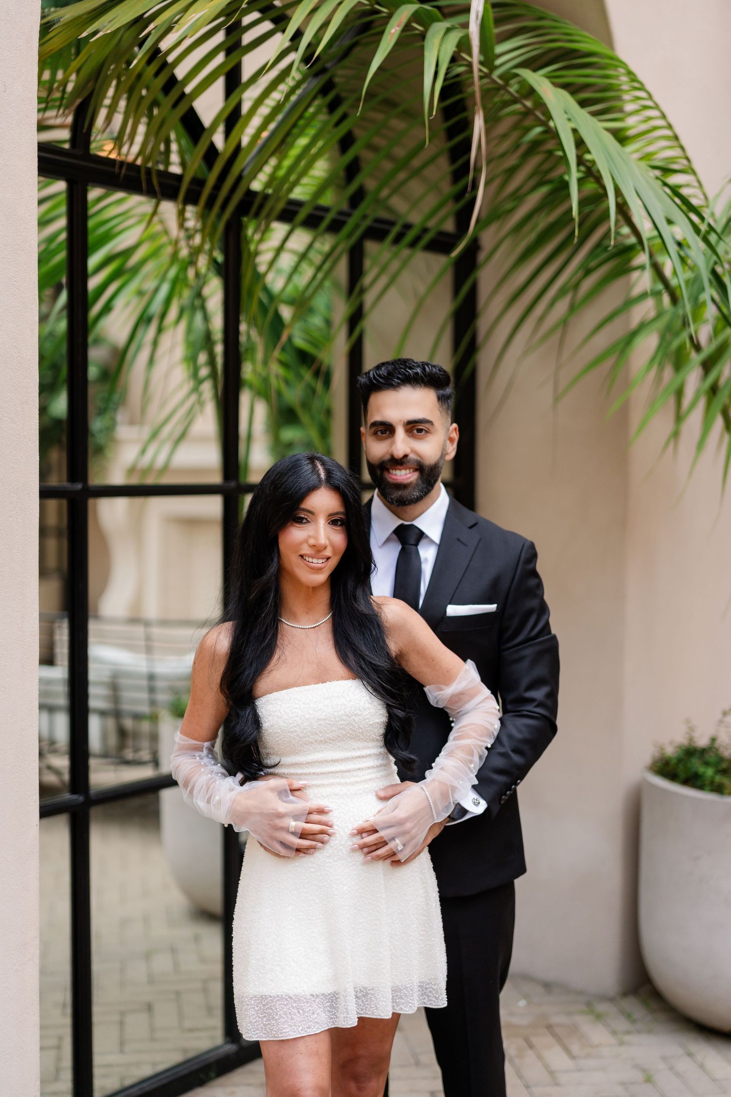 Romantic couple embracing in courtyard with palm trees and arched windows.