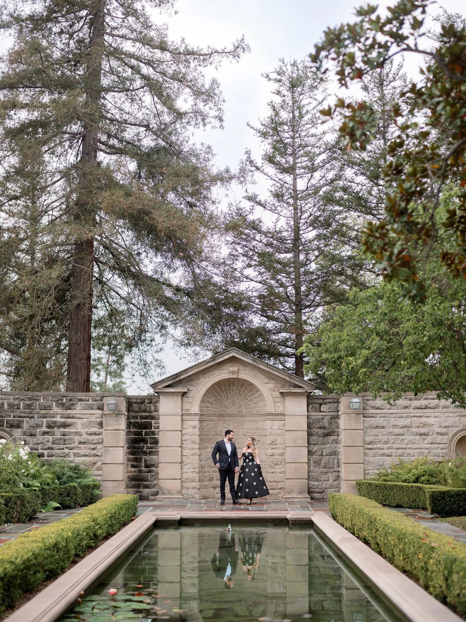 A couple poses by a reflecting pool with formal gardens and stone architecture.