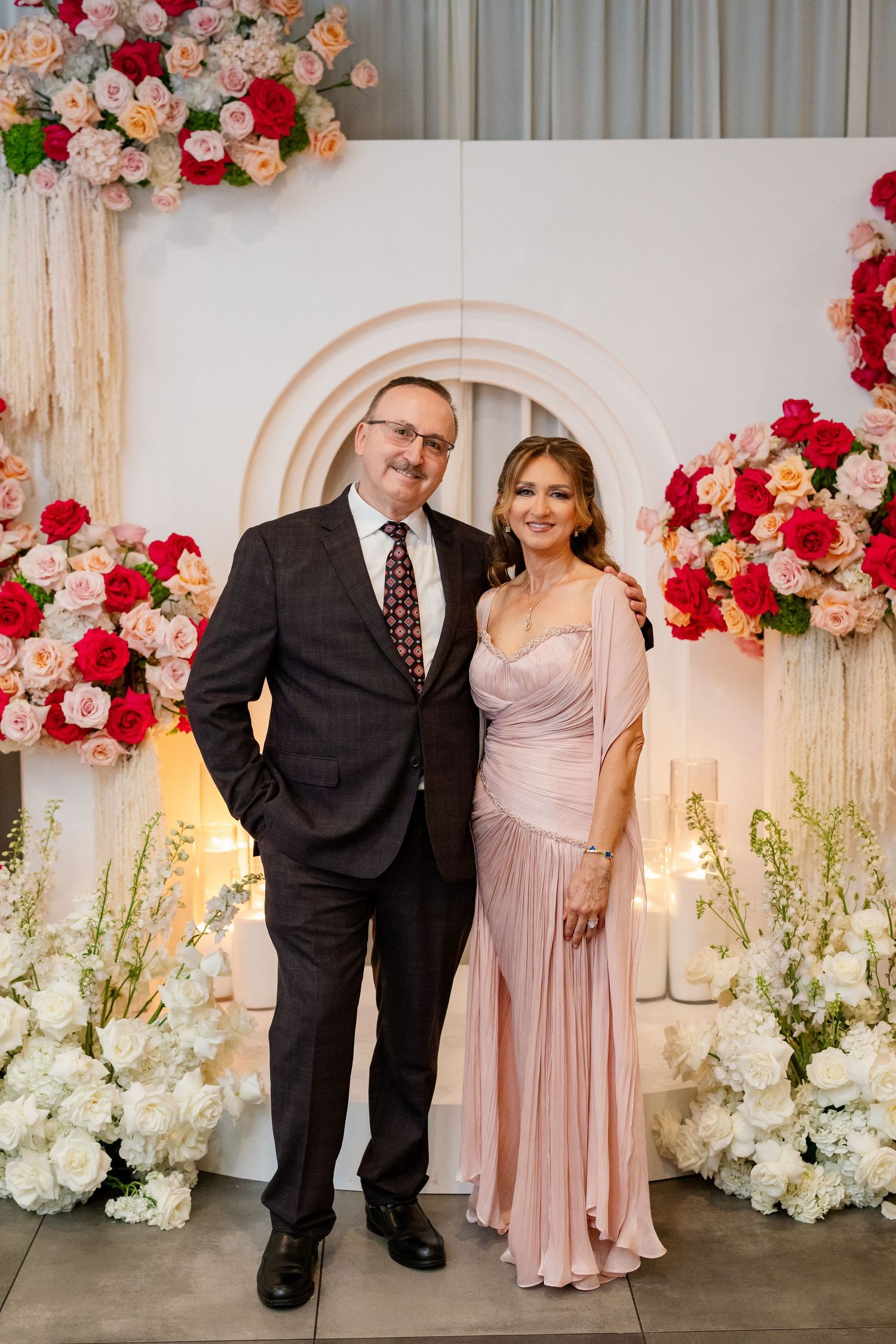 Couple posing together at a floral wedding backdrop with pink and red roses, man in suit and woman in pink gown.