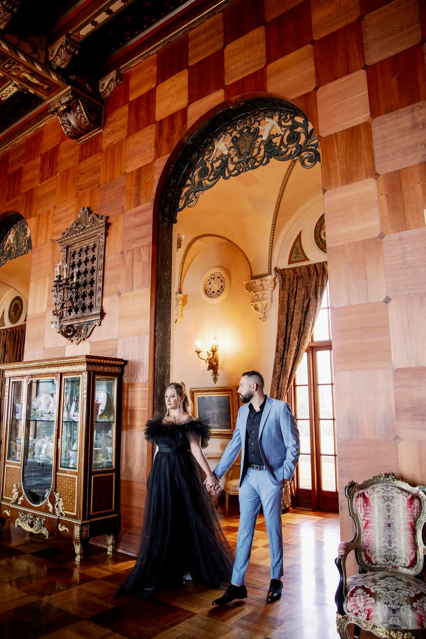 A couple stands in an ornate room with checkered walls and antique furnishings.