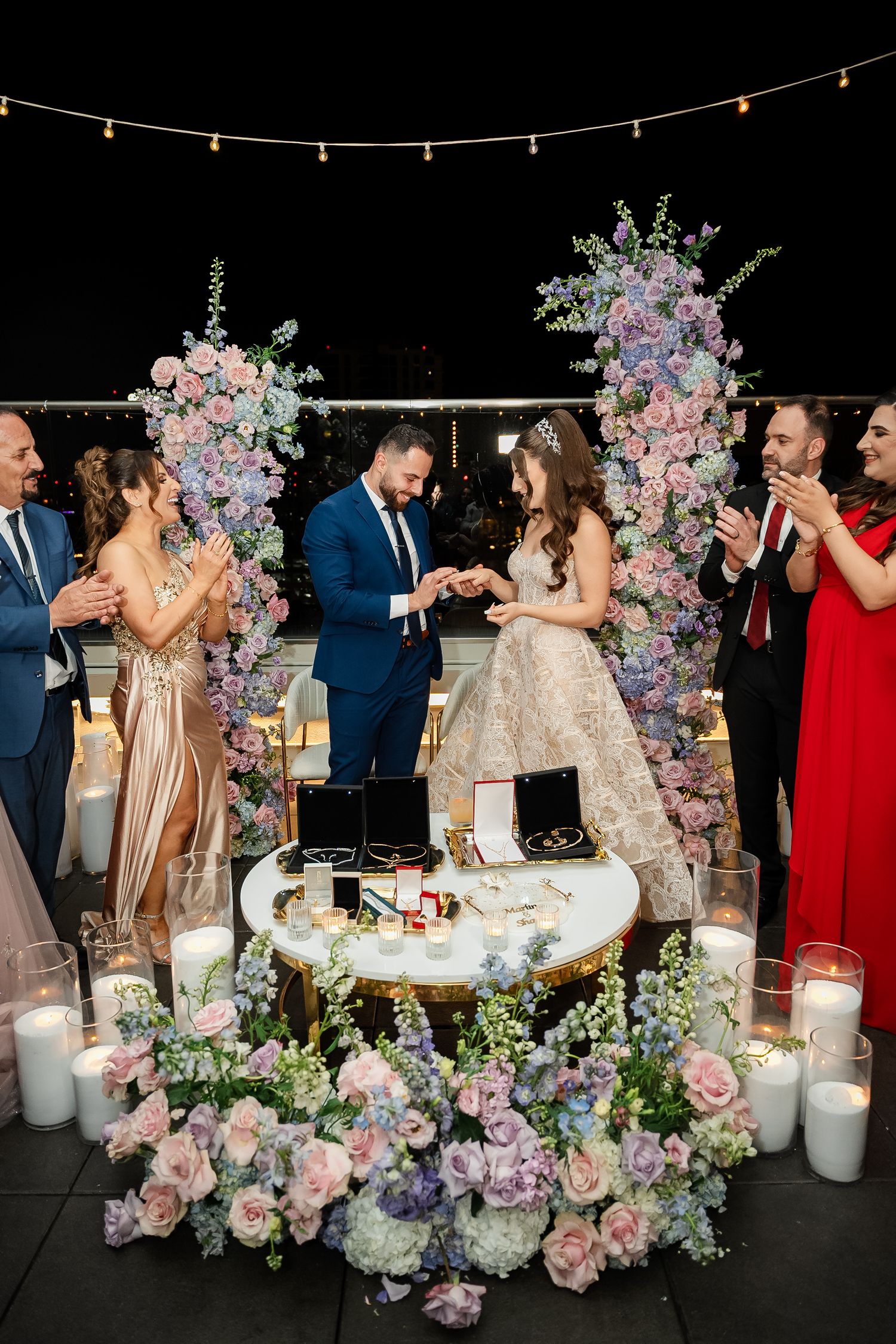 Wedding reception features elegant table settings with pink and purple floral arrangements and string lights.