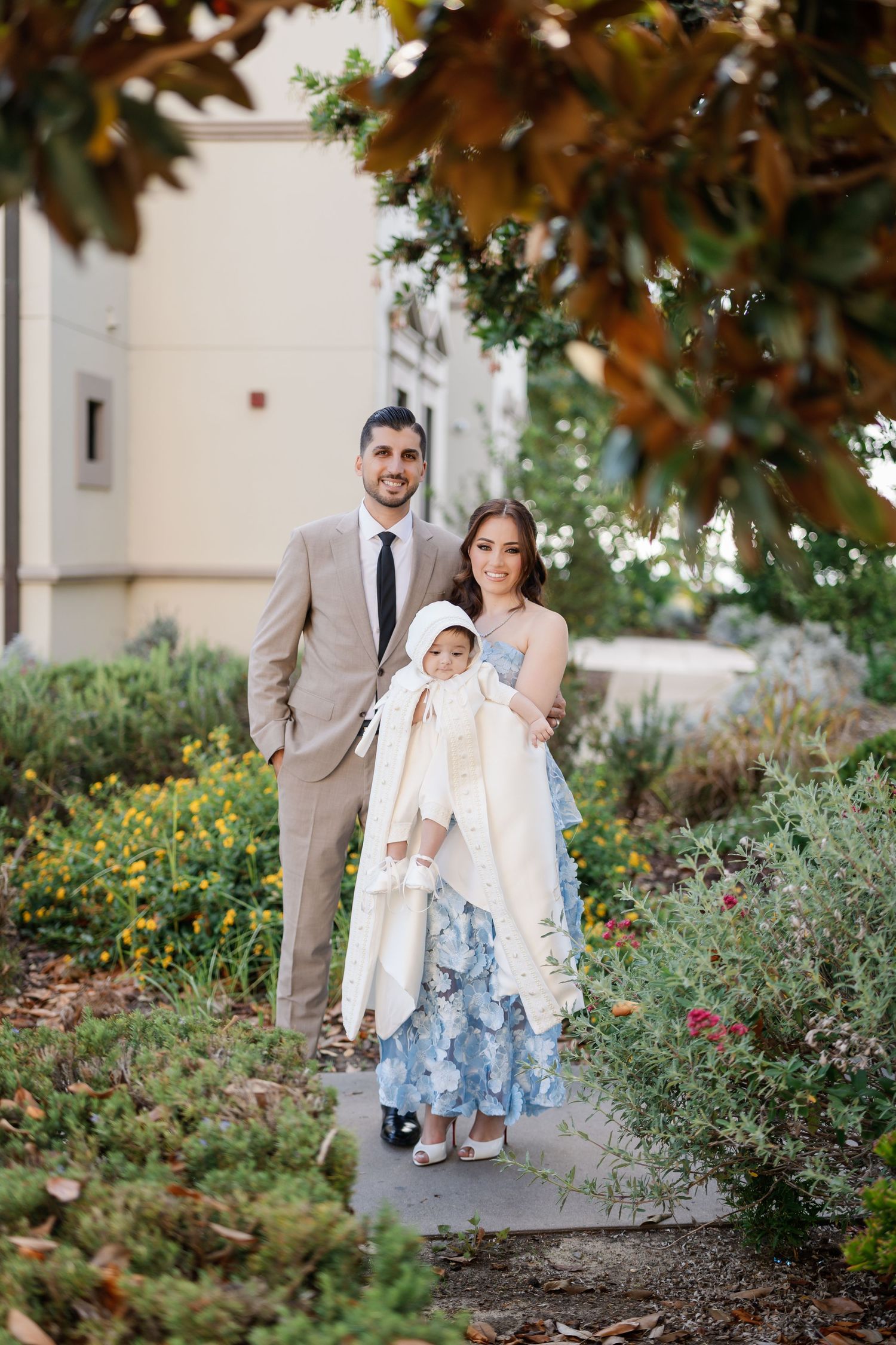 A family in formal attire poses together in a garden setting with blooming flowers and greenery.