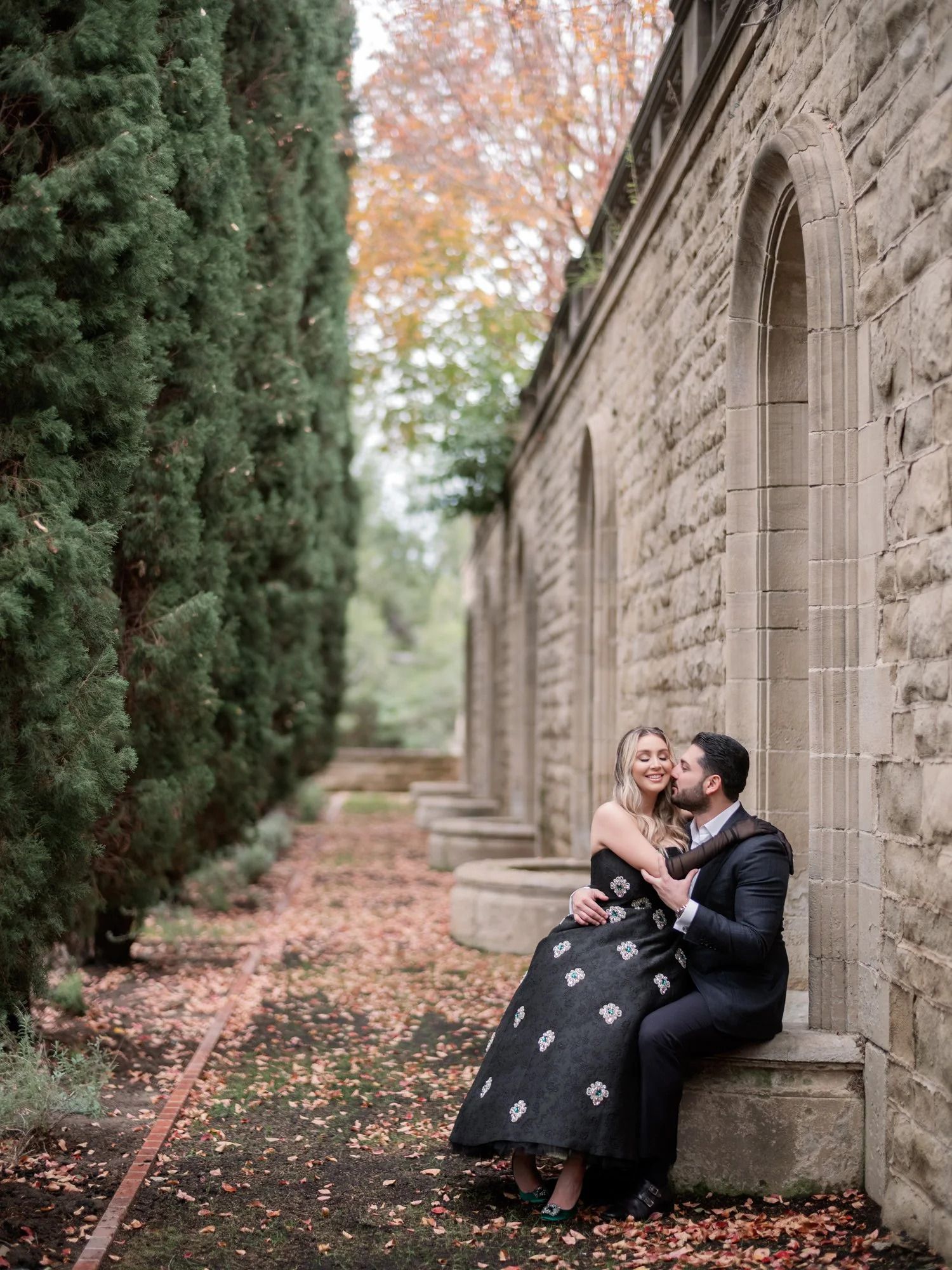 A couple shares an intimate moment on stone steps surrounded by autumn foliage and cypress trees.