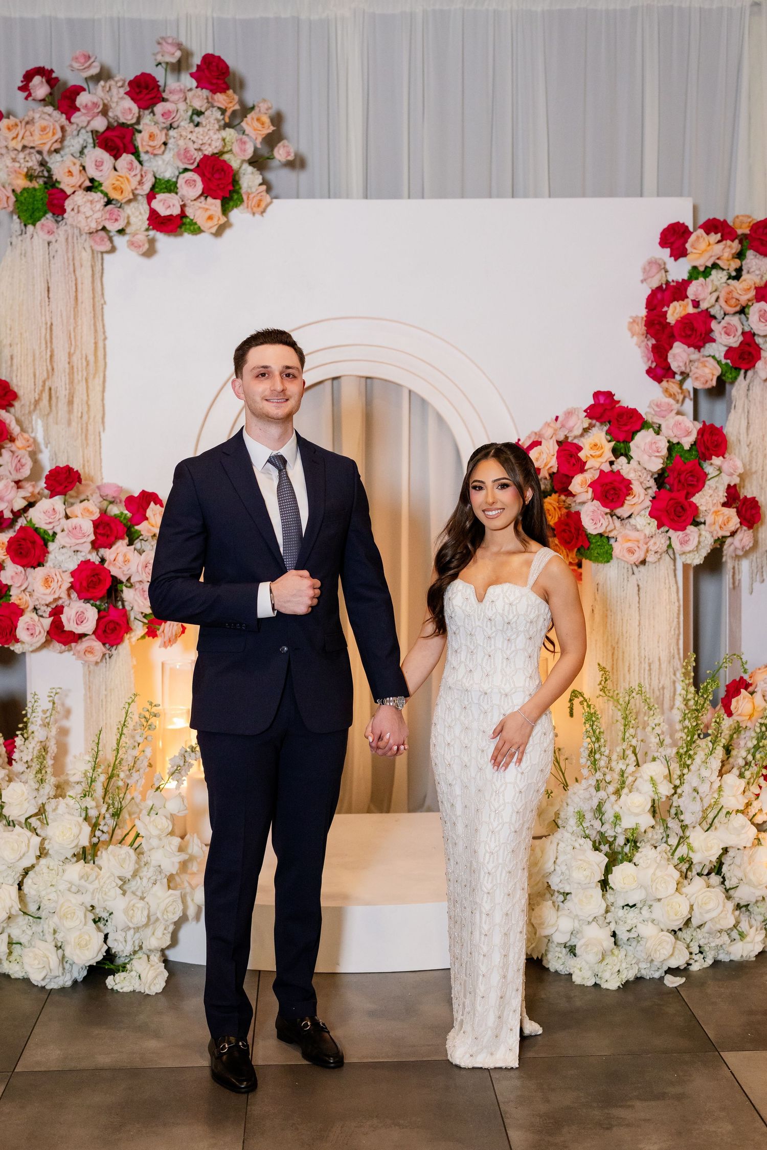 Couple in formal attire posing before a floral arch backdrop with pink, red, and white roses at an elegant event.