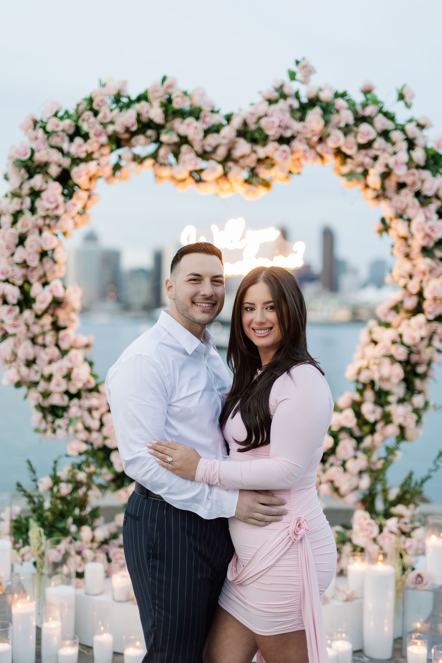 Couple celebrating engagement under pink floral arch with city skyline and candlelit setting.
