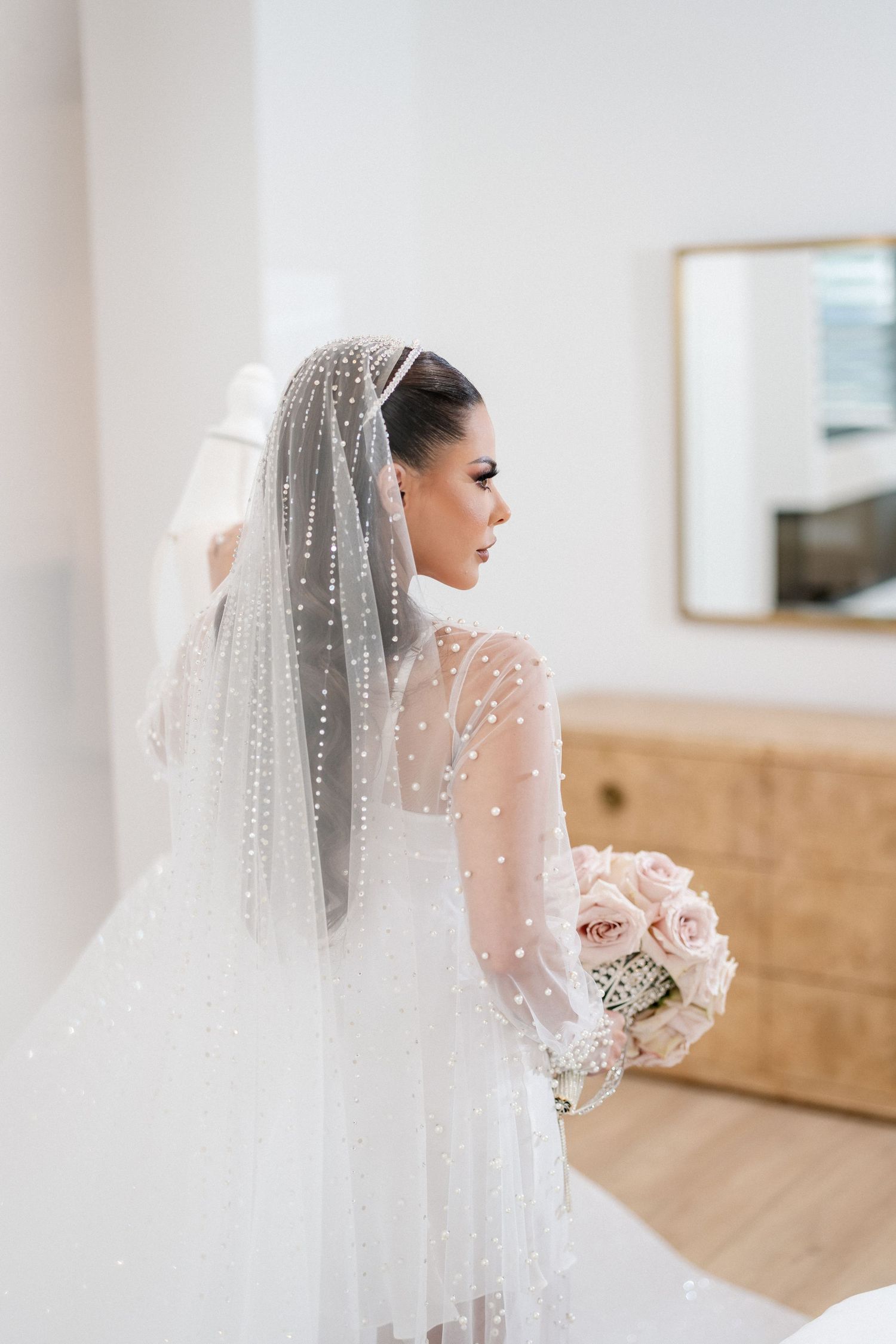 A bride in a sheer long-sleeved gown with a flowing veil poses in an elegant indoor setting.