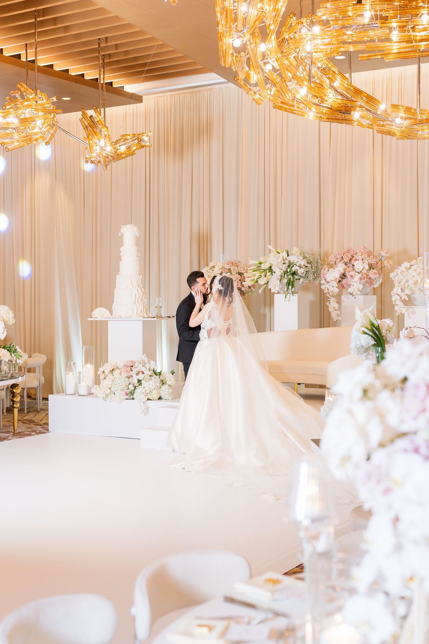 Elegant wedding reception hall features golden chandeliers and white table settings with floral centerpieces.