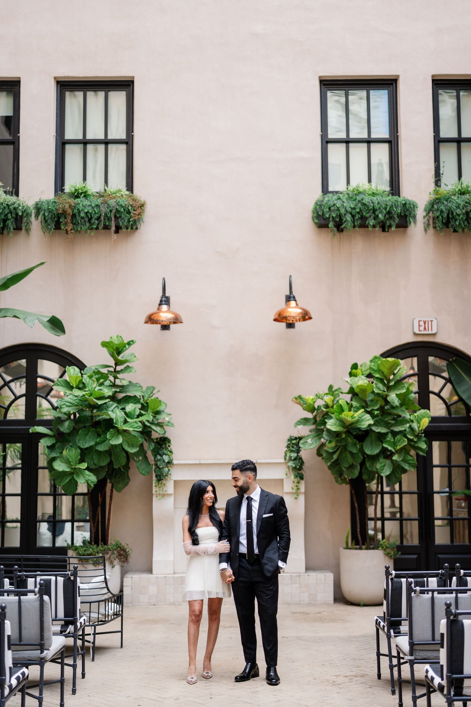 Elegant couple standing in outdoor courtyard with beige walls and lush greenery.
