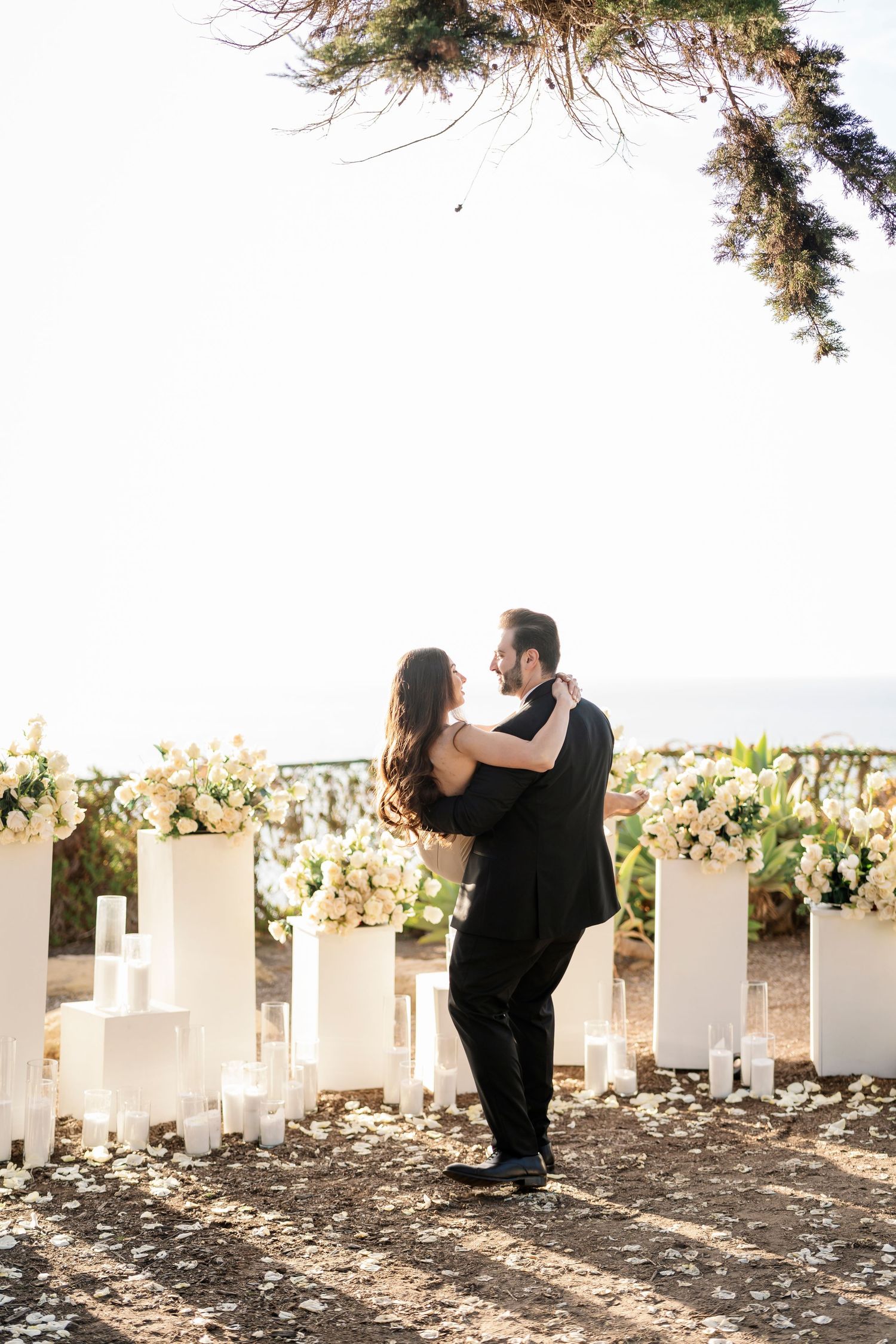 Romantic couple embraces near white pillars decorated with flowers in outdoor setting.