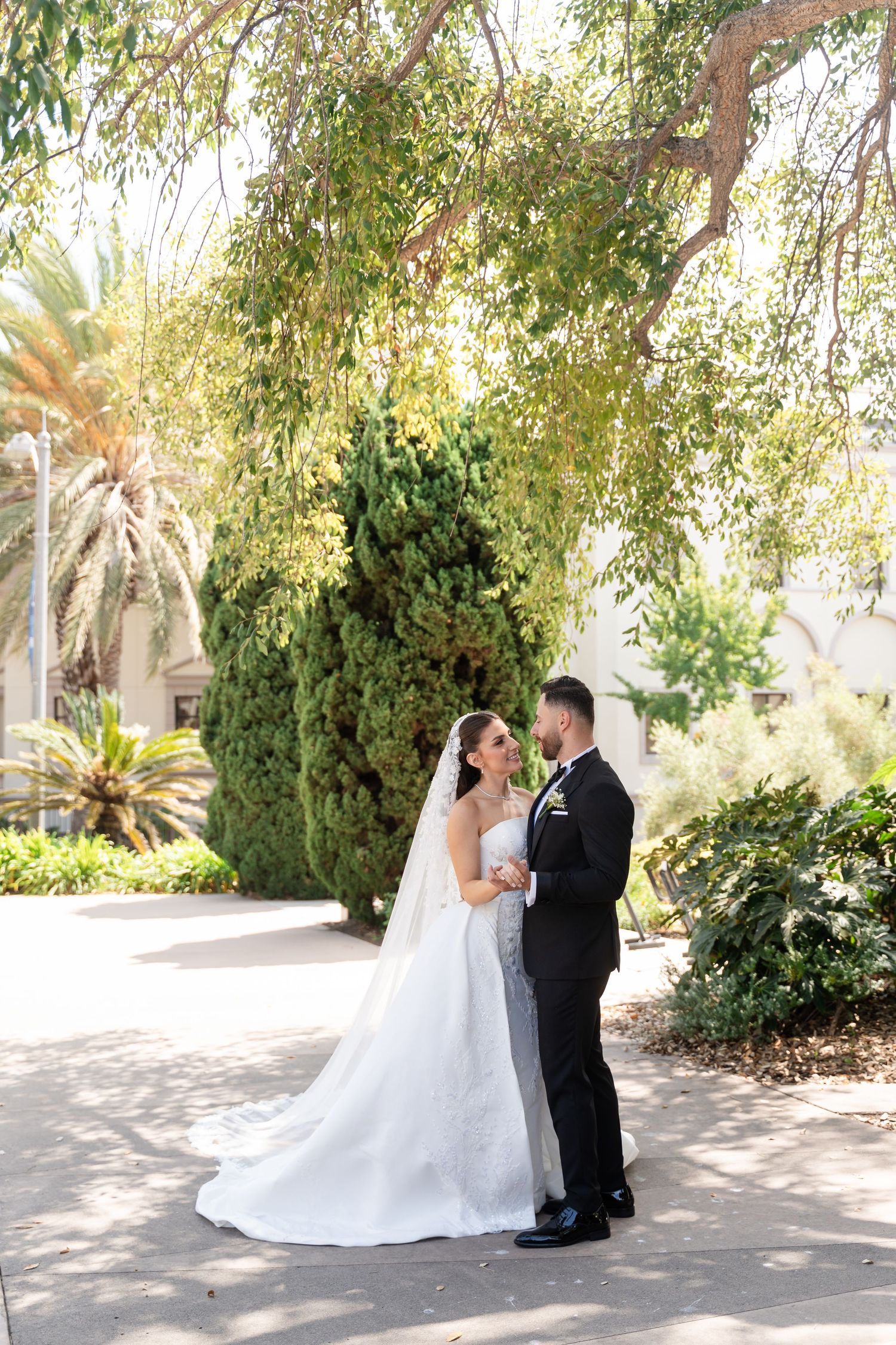 A romantic wedding portrait of a couple embracing on a garden path with palm trees and greenery in the background.