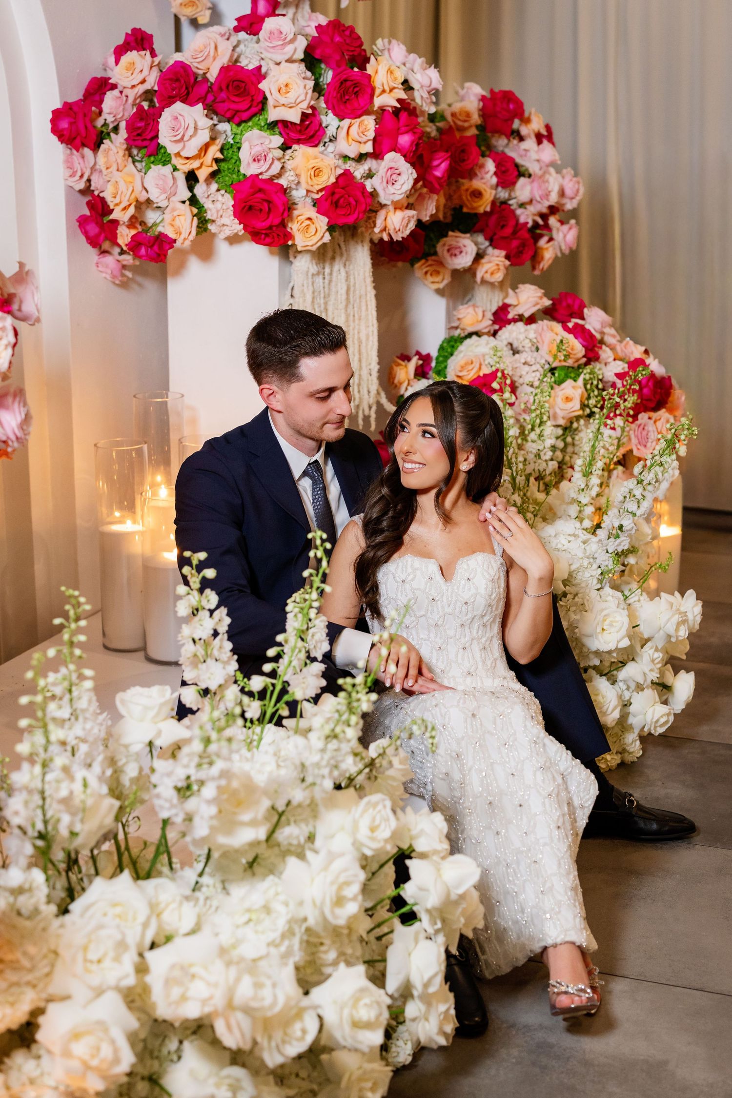 Bride and groom sit surrounded by lush white and pink floral arrangements with candles at their romantic wedding reception.