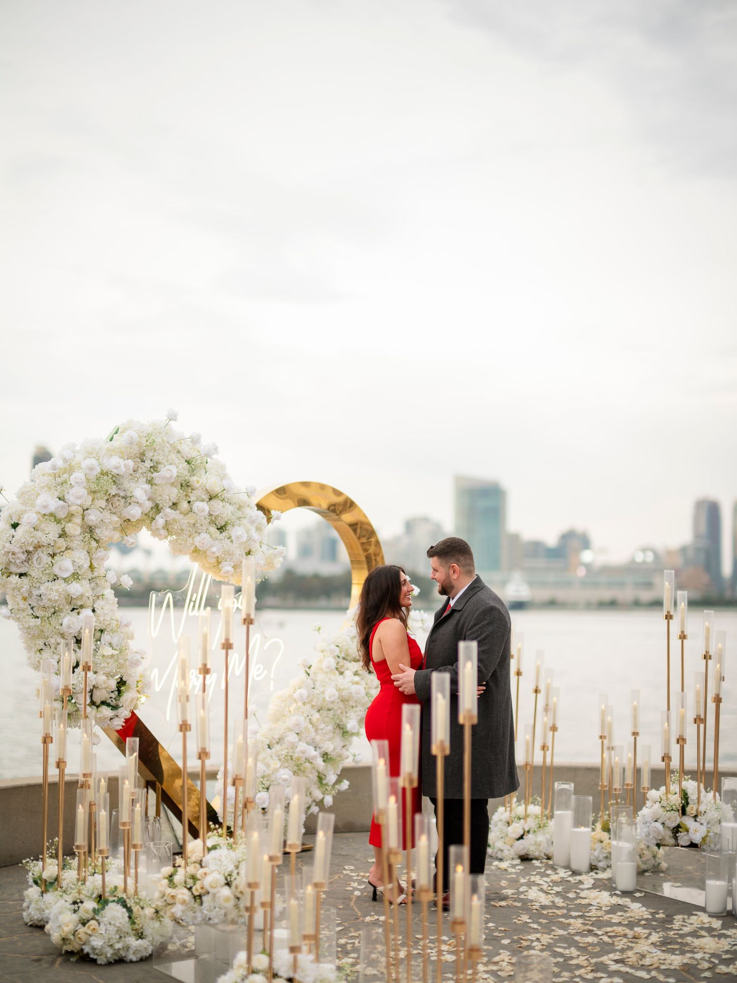 An elegant waterfront proposal scene with white floral arrangements and golden candle holders.