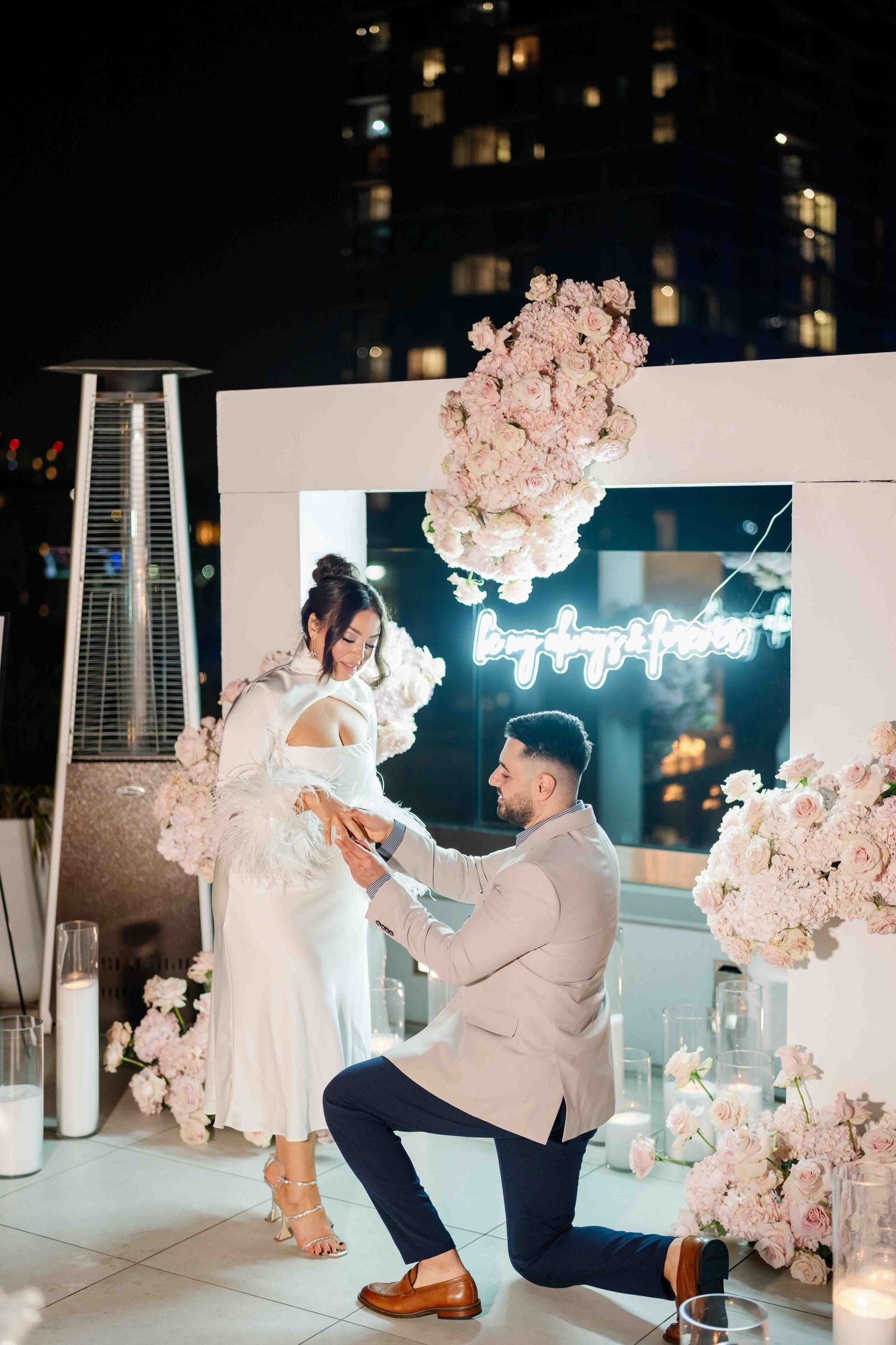 Nighttime rooftop proposal scene with pink floral arrangements and ambient lighting.