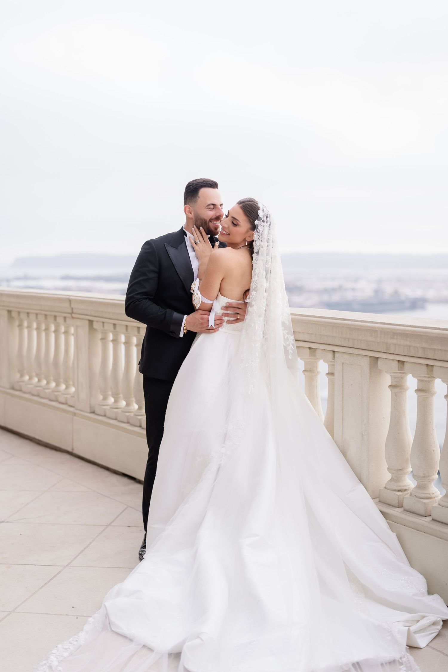 Newlyweds share an intimate moment on a balcony overlooking a city landscape.