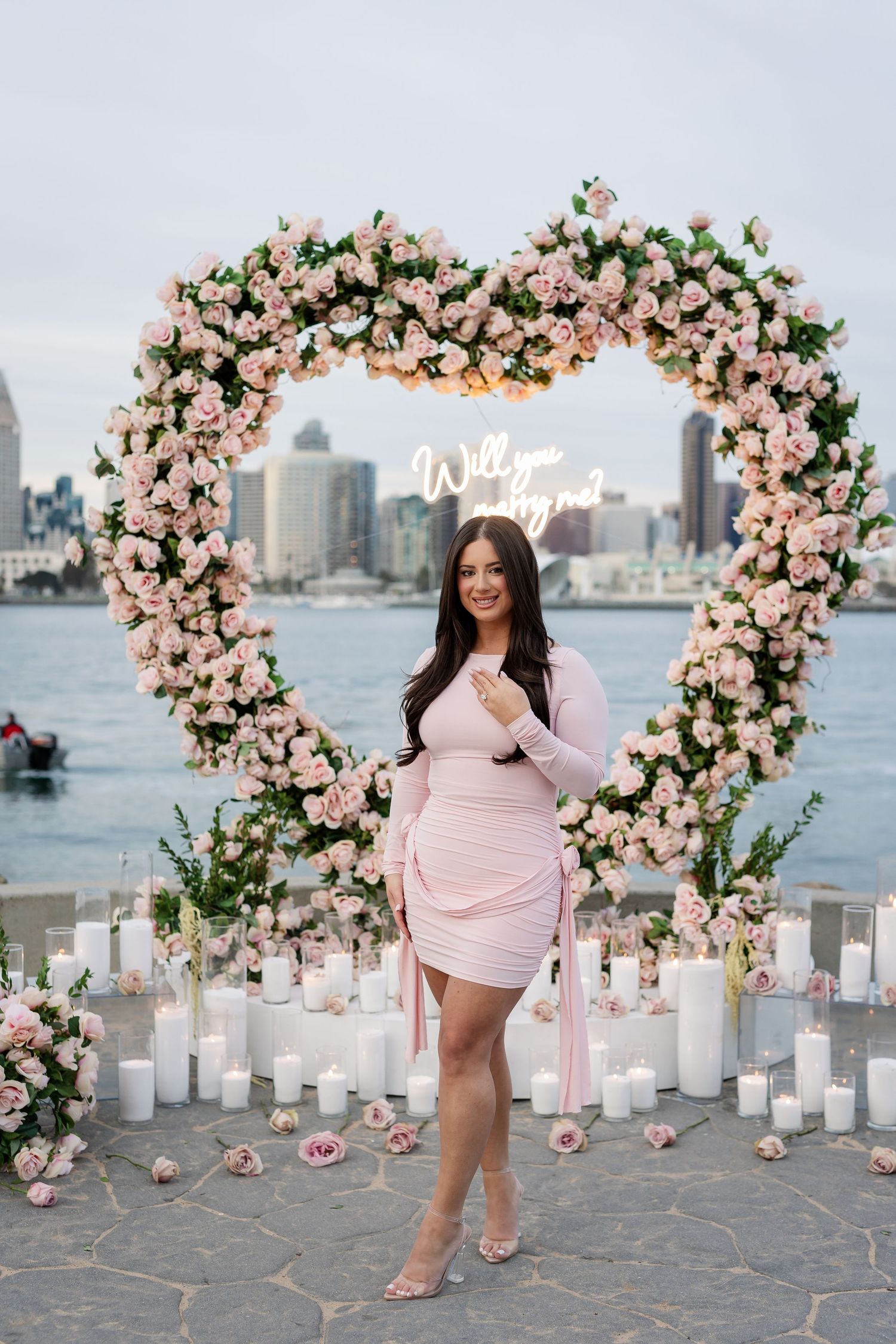 Woman in pink dress poses by floral arch with cityscape and candles creating romantic ambiance.