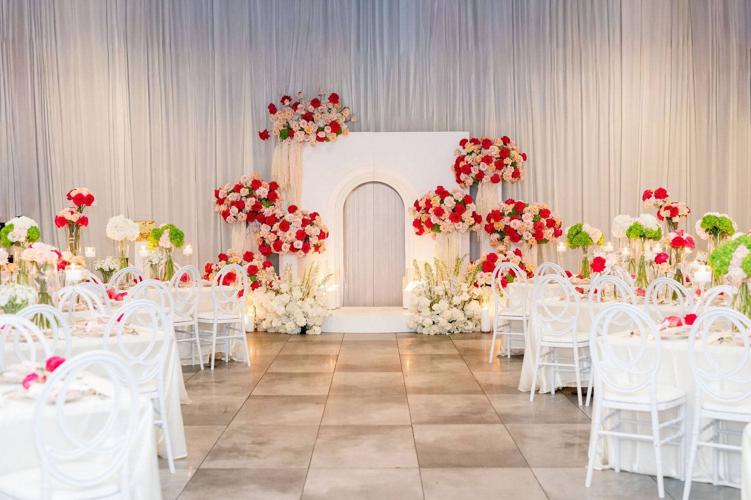 Elegant wedding reception hall with white chairs, red and white floral arrangements, and a decorative arch backdrop.