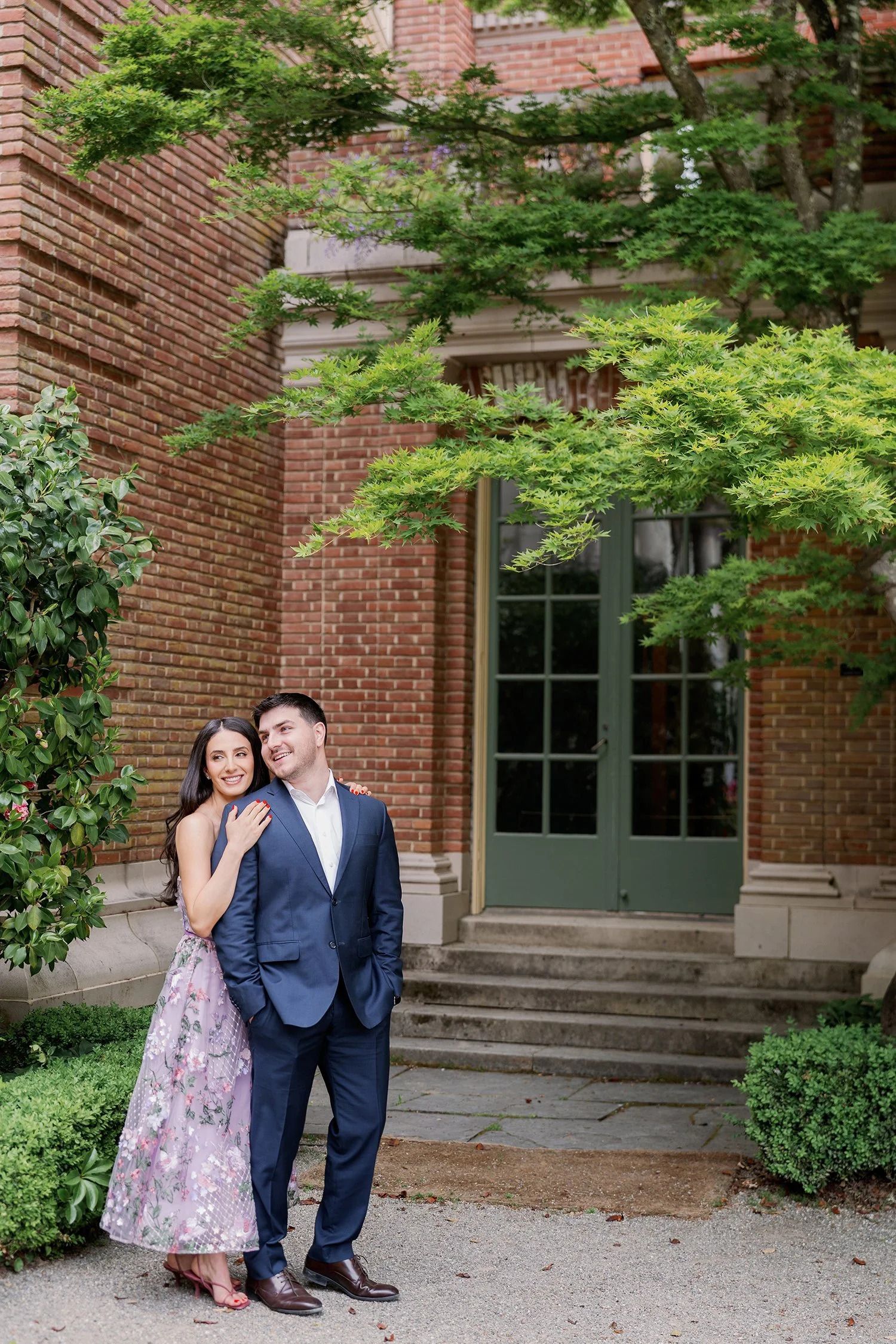 A couple poses together against a brick building surrounded by lush green foliage.