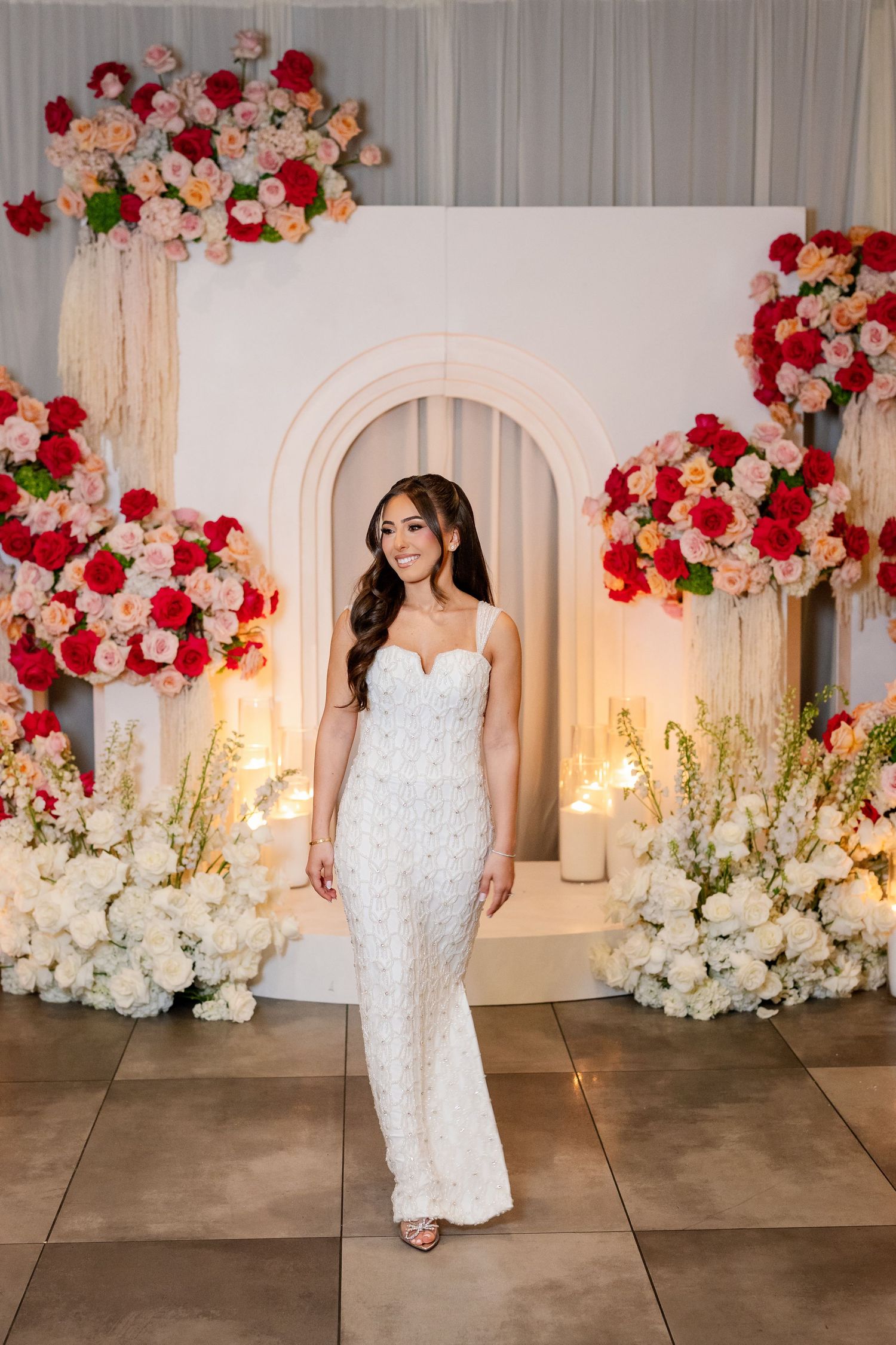 Bride in white lace gown poses before floral arch backdrop with red and pink roses and candles.