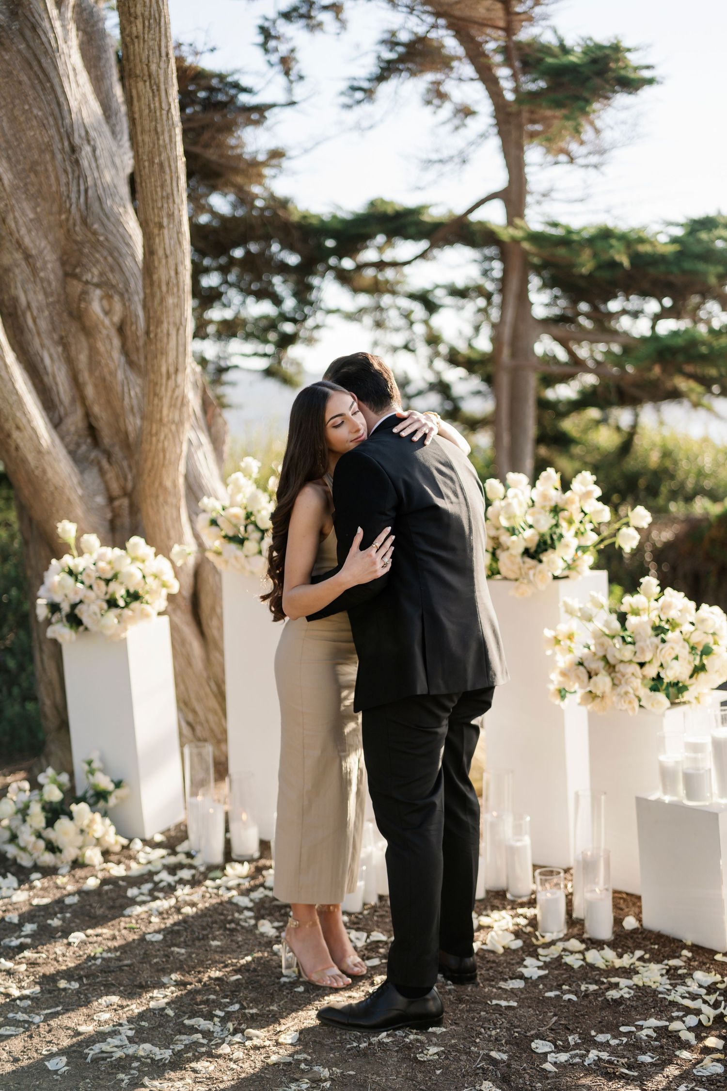 Couple embraces among white pillars decorated with cream flowers in outdoor garden setting.